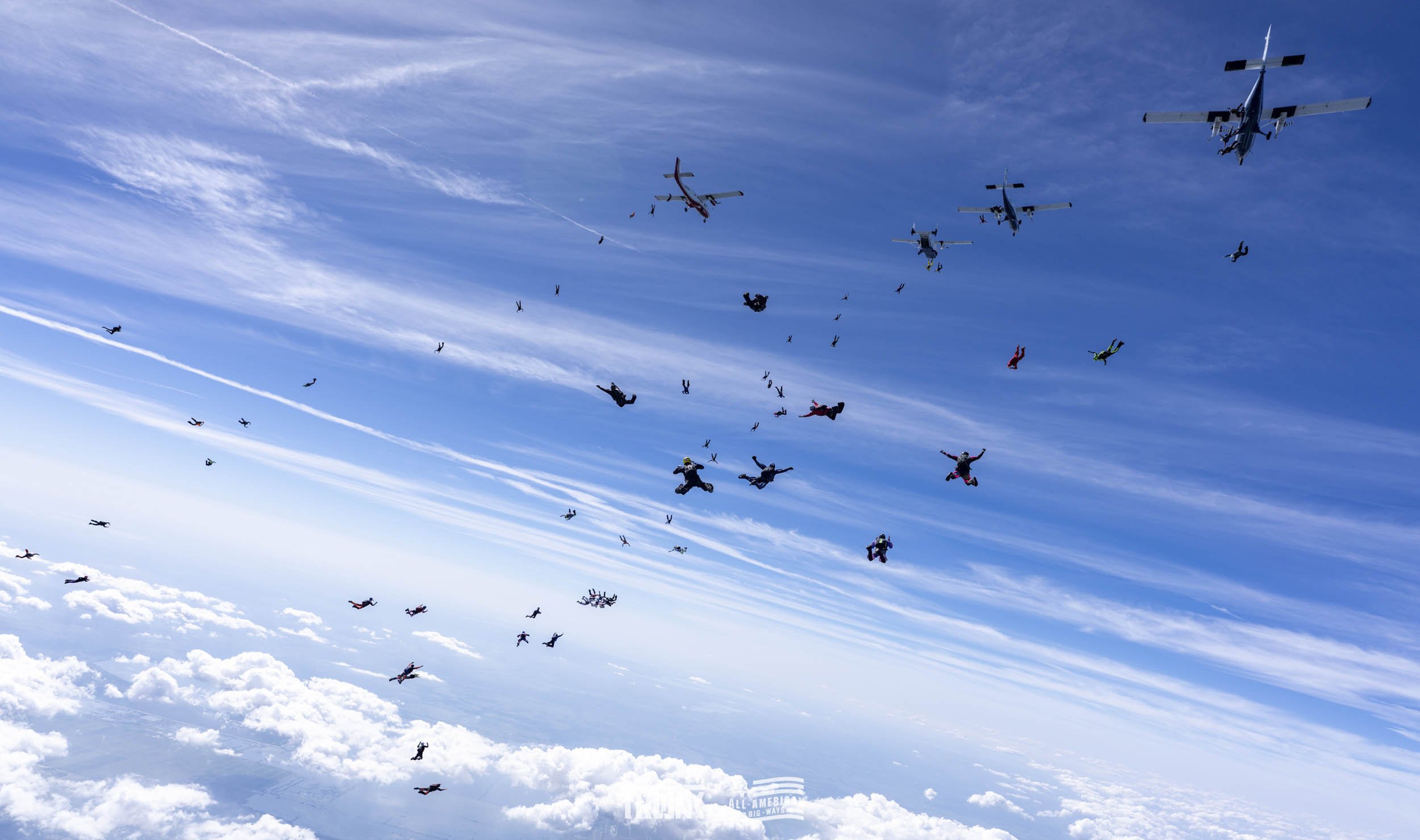 Skydivers falling through the sky with clouds below and several airplanes above, captured during free fall.