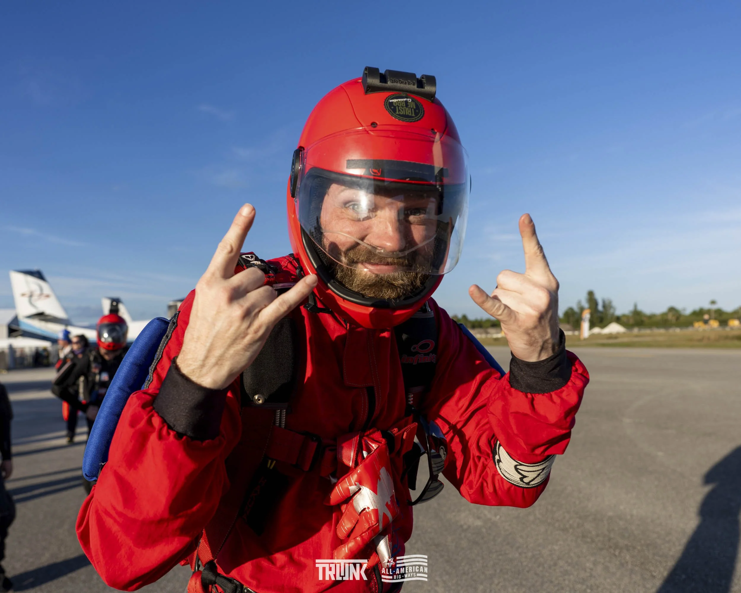 A man in a red racing suit and helmet making the rock on hand gesture at an airfield.