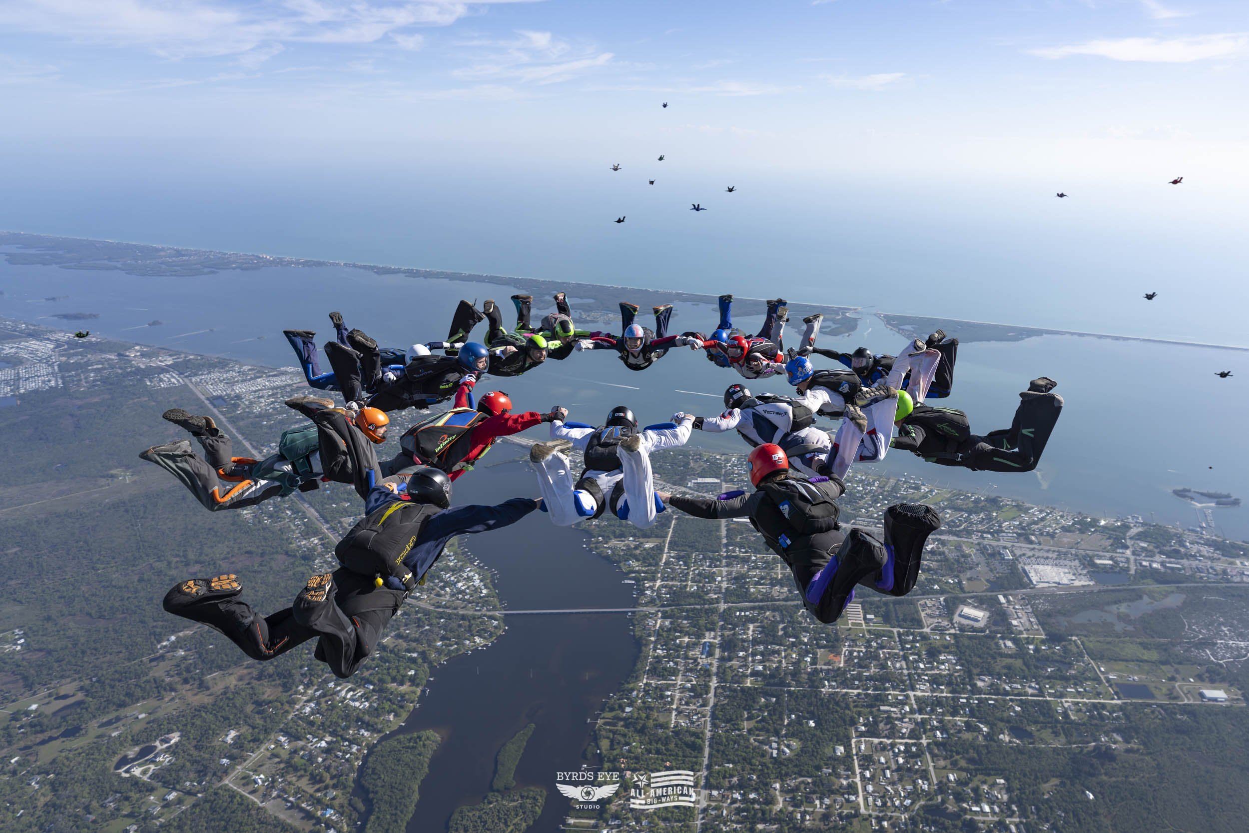 A group of skydivers in freefall during a jump, holding hands in a circle over a landscape with water, forests, and a city, with other skydivers visible in the background.