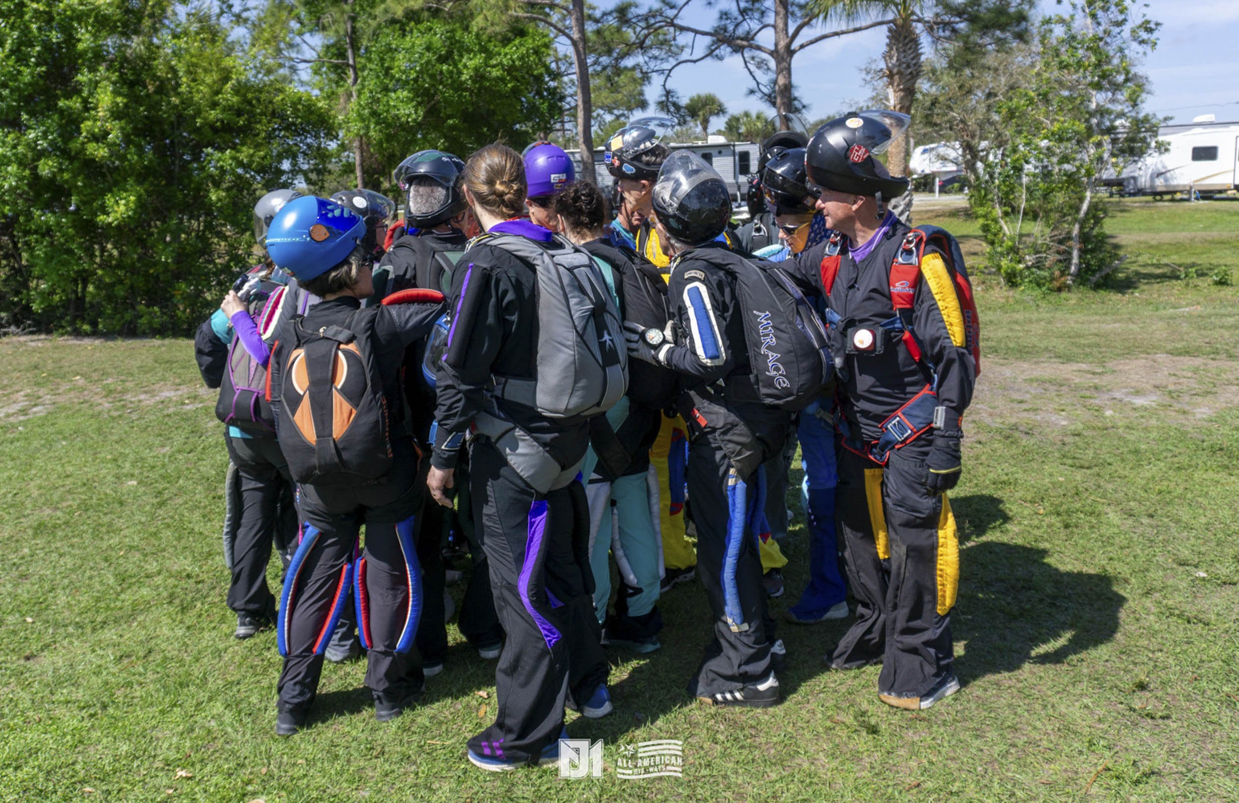 Group of skydivers wearing helmets and jumpsuits, gathered outdoors on grass.