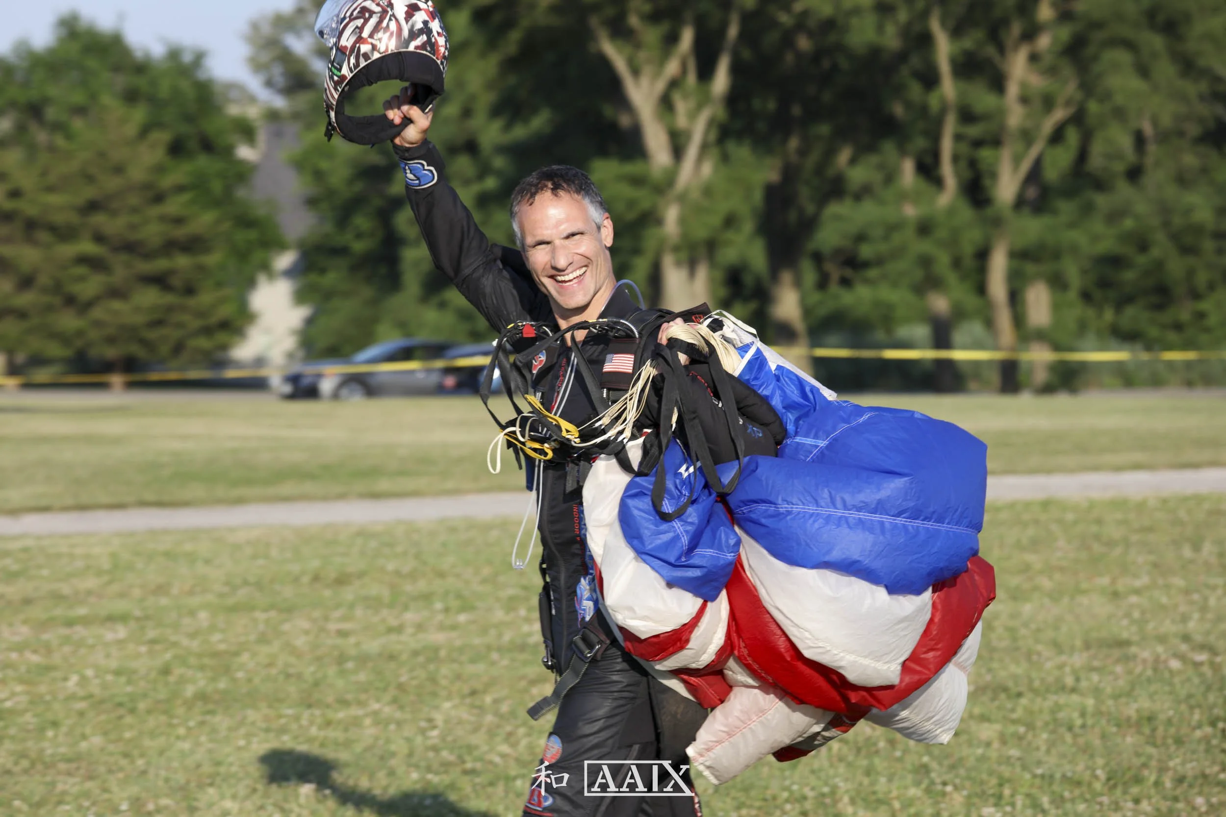 A smiling man in skydiving gear holding a helmet in one hand, standing outdoors on grass with trees in the background.