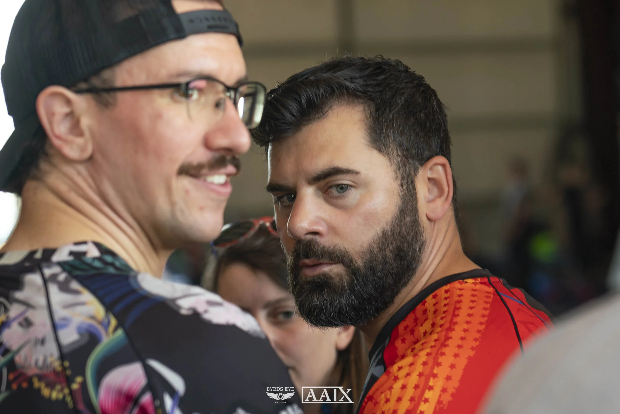 Two men with serious expressions looking towards the camera, one with a beard and dark hair wearing a red and black shirt, and the other with glasses, a mustache, and a backwards cap wearing a colorful shirt, in an indoor setting.