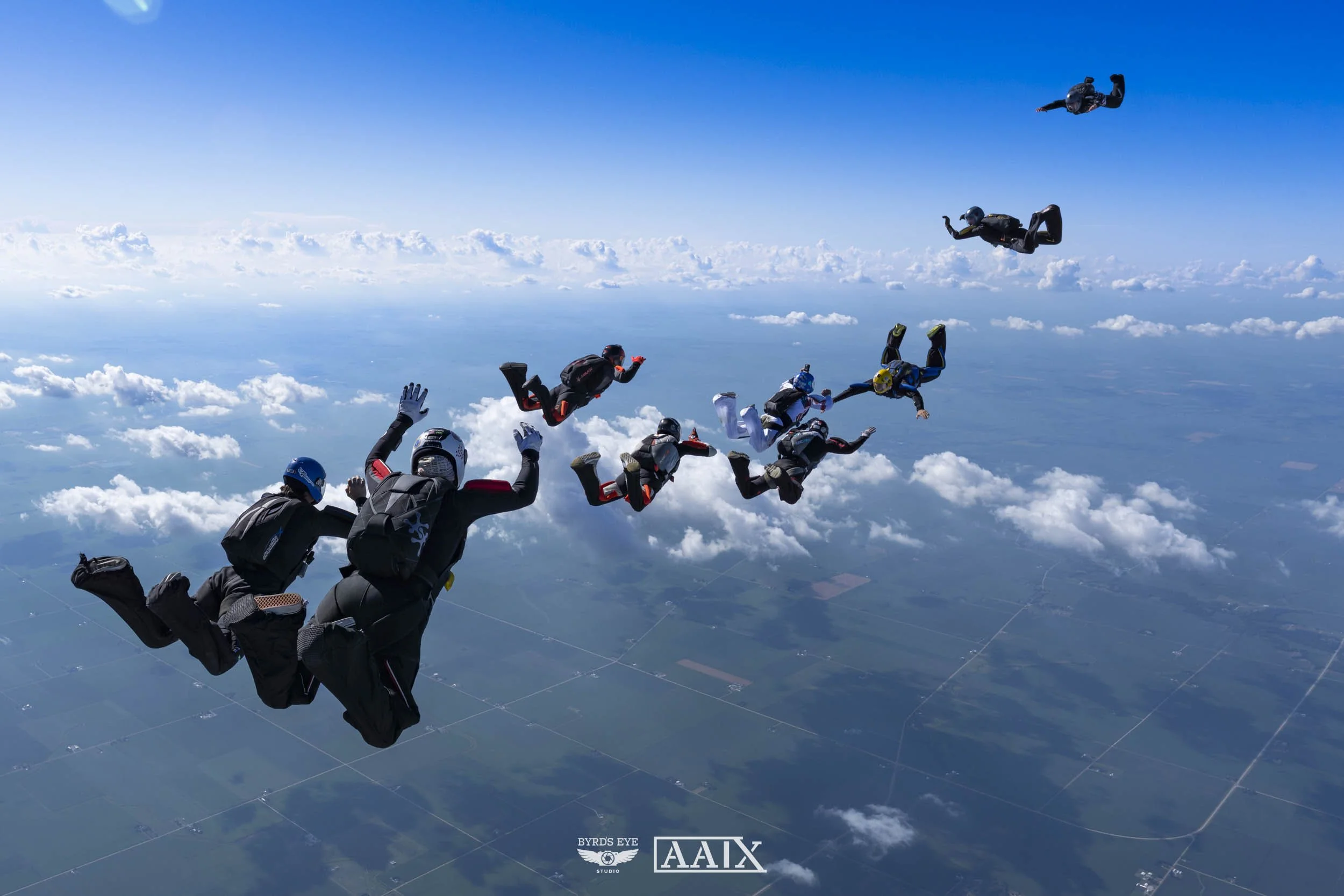 A group of skydivers free-falling through the sky over a landscape of clouds and farmland, with some skydivers in the foreground and others in the background.