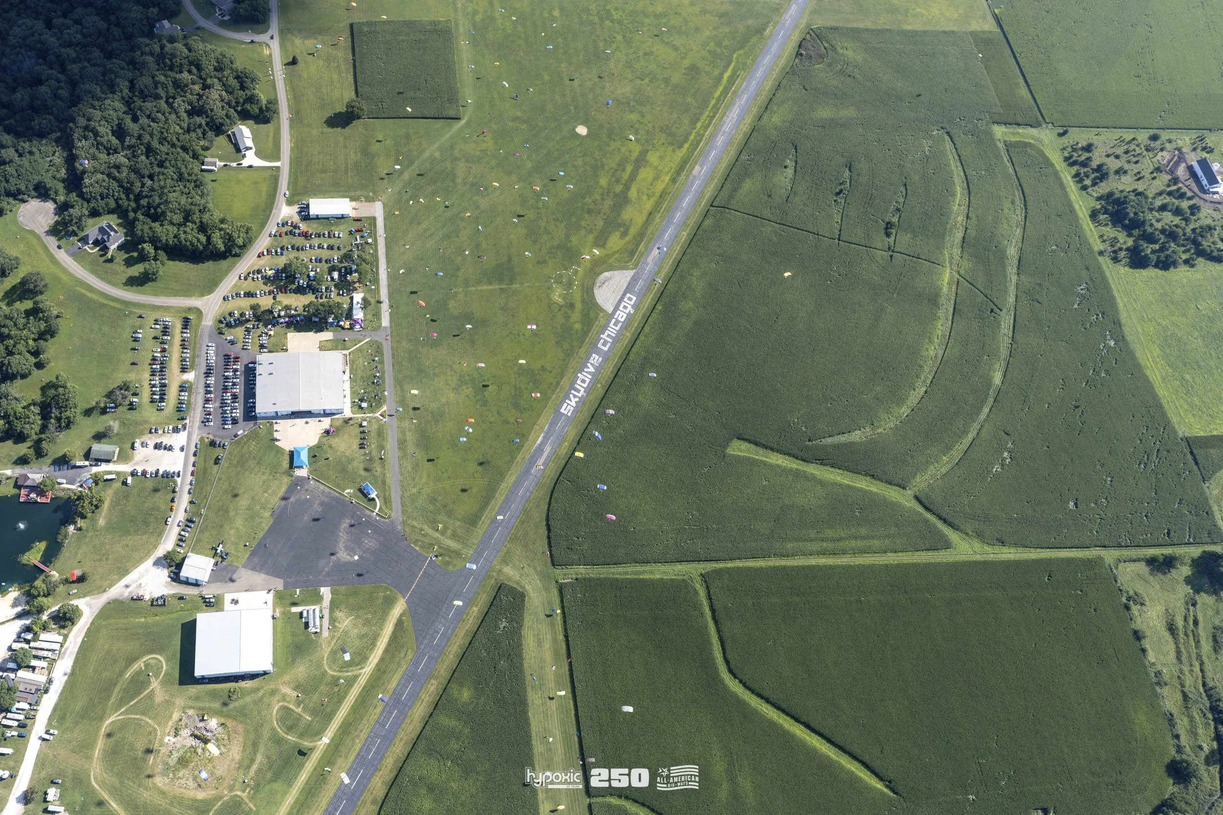 Aerial view of Skydive Chicago, an outdoor skydiving facility with a large parking lot, a clubhouse, and open fields, surrounded by farmland and trees.