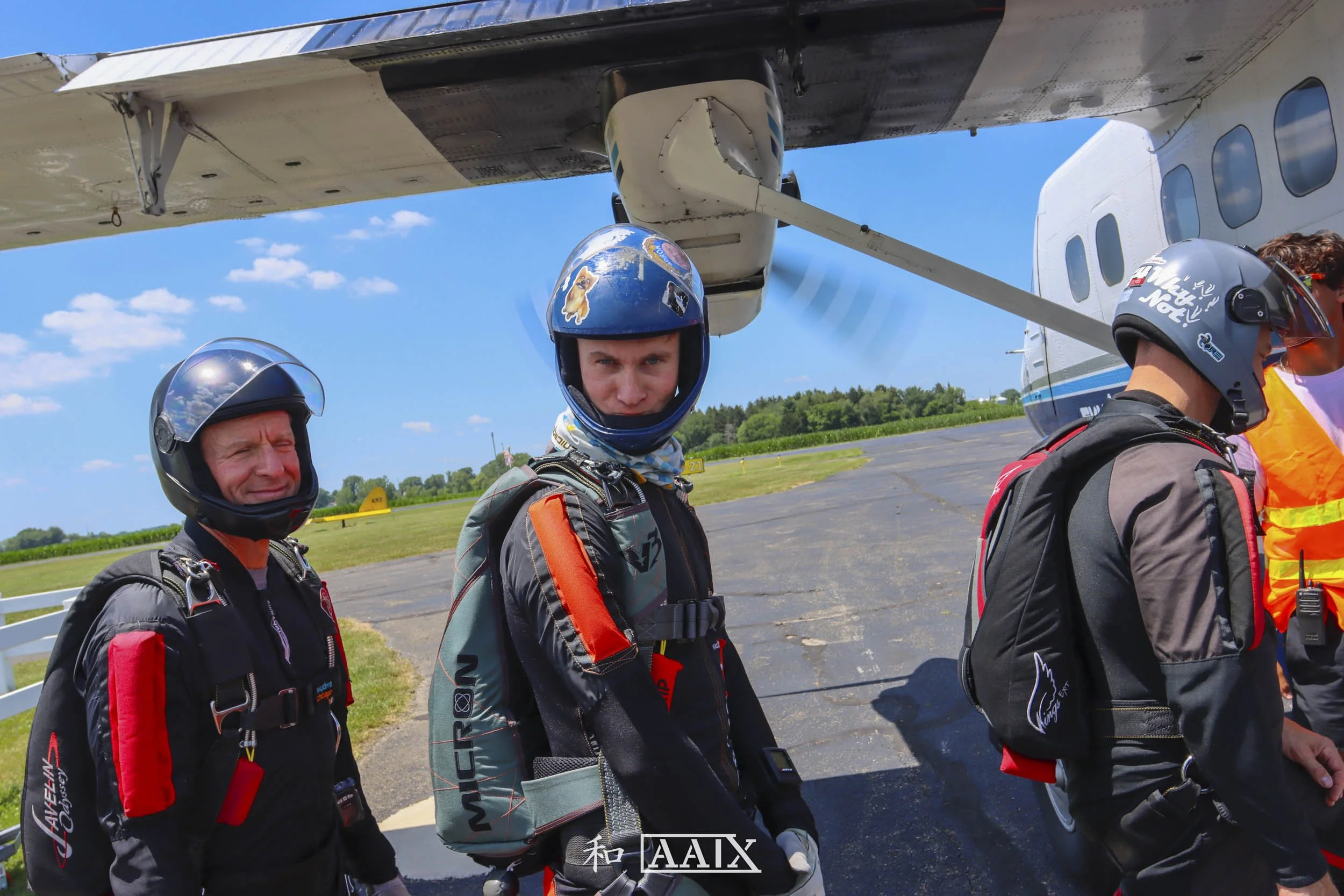 Group of skydivers in flight suits and helmets preparing for jump, standing near an aircraft on the tarmac.