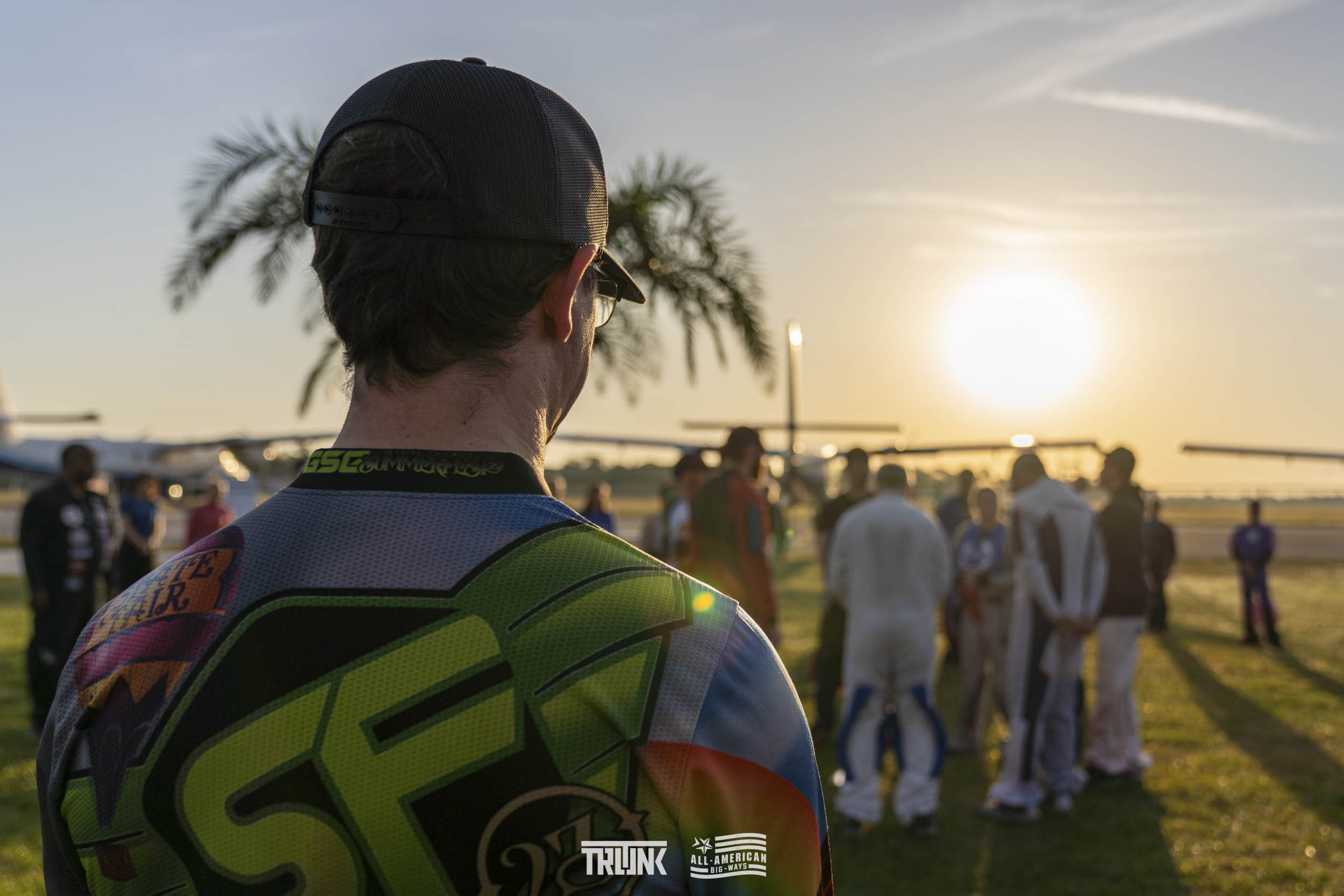 A man wearing a racing suit and baseball cap stands in the foreground at an outdoor racing event during sunset, with planes and a group of people in racing suits in the background.