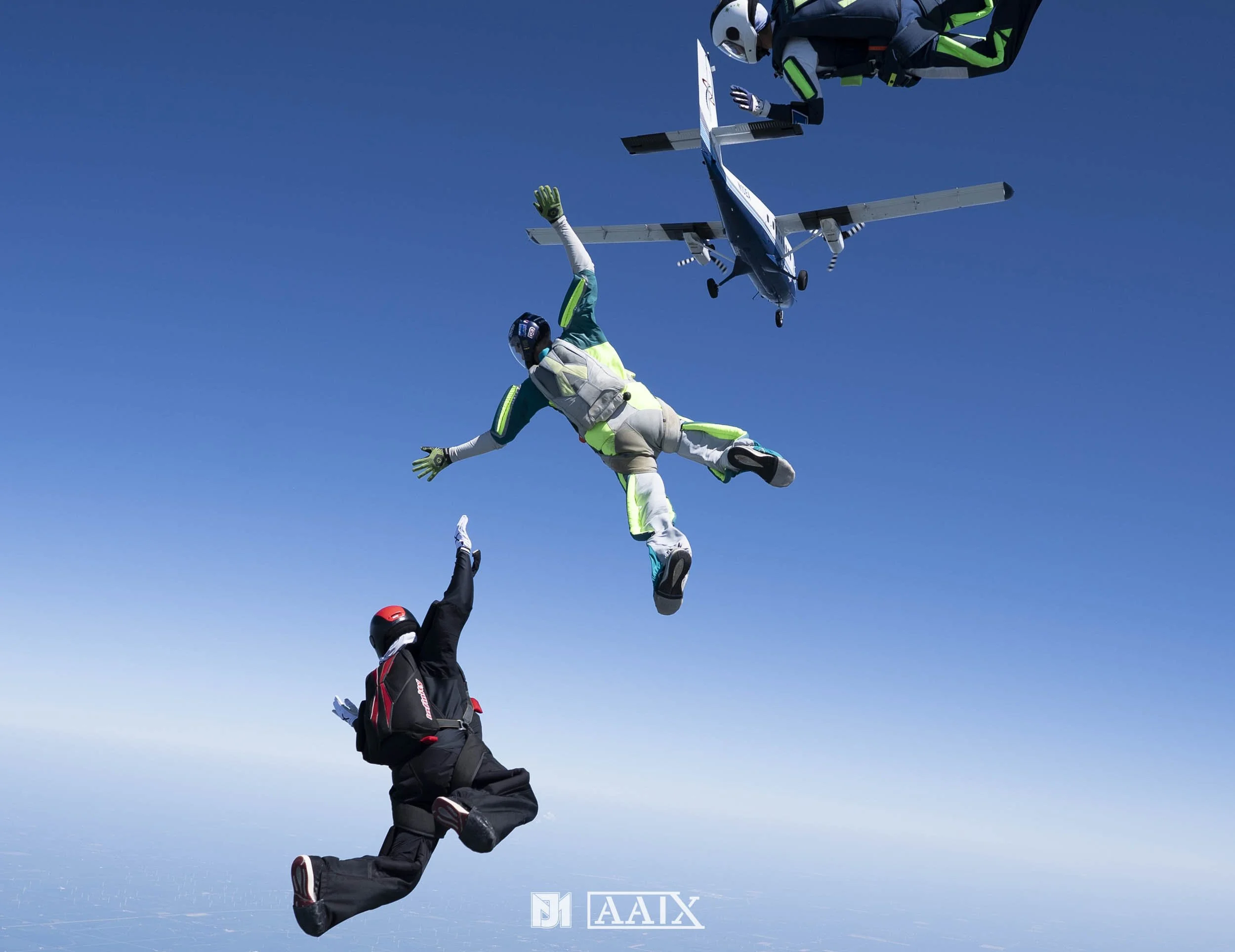 Skydivers mid-air in jumpsuits and helmets with an airplane in the background against a clear blue sky.
