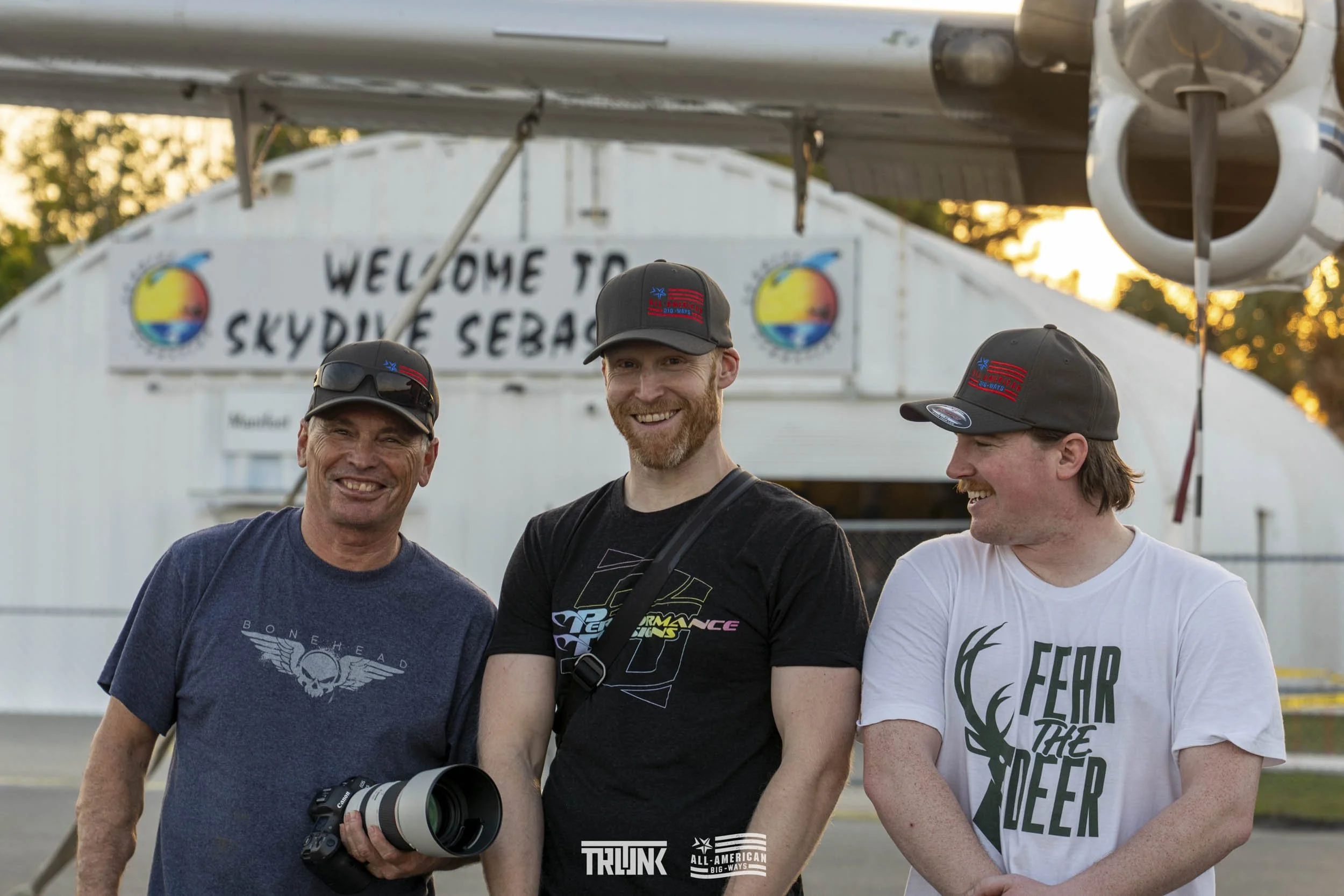 Three men smiling in front of a skydive sign, with a plane overhead; one man holds a camera with a large telephoto lens.