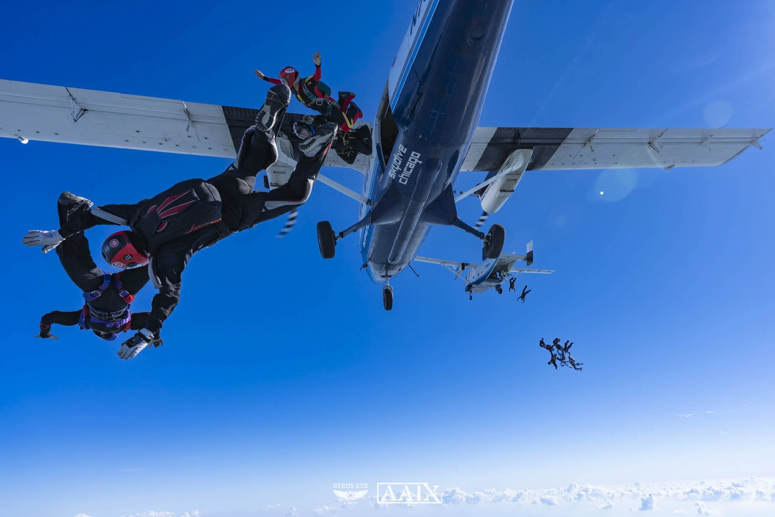 Skydivers jumping out of a large airplane with blue sky and clouds below.