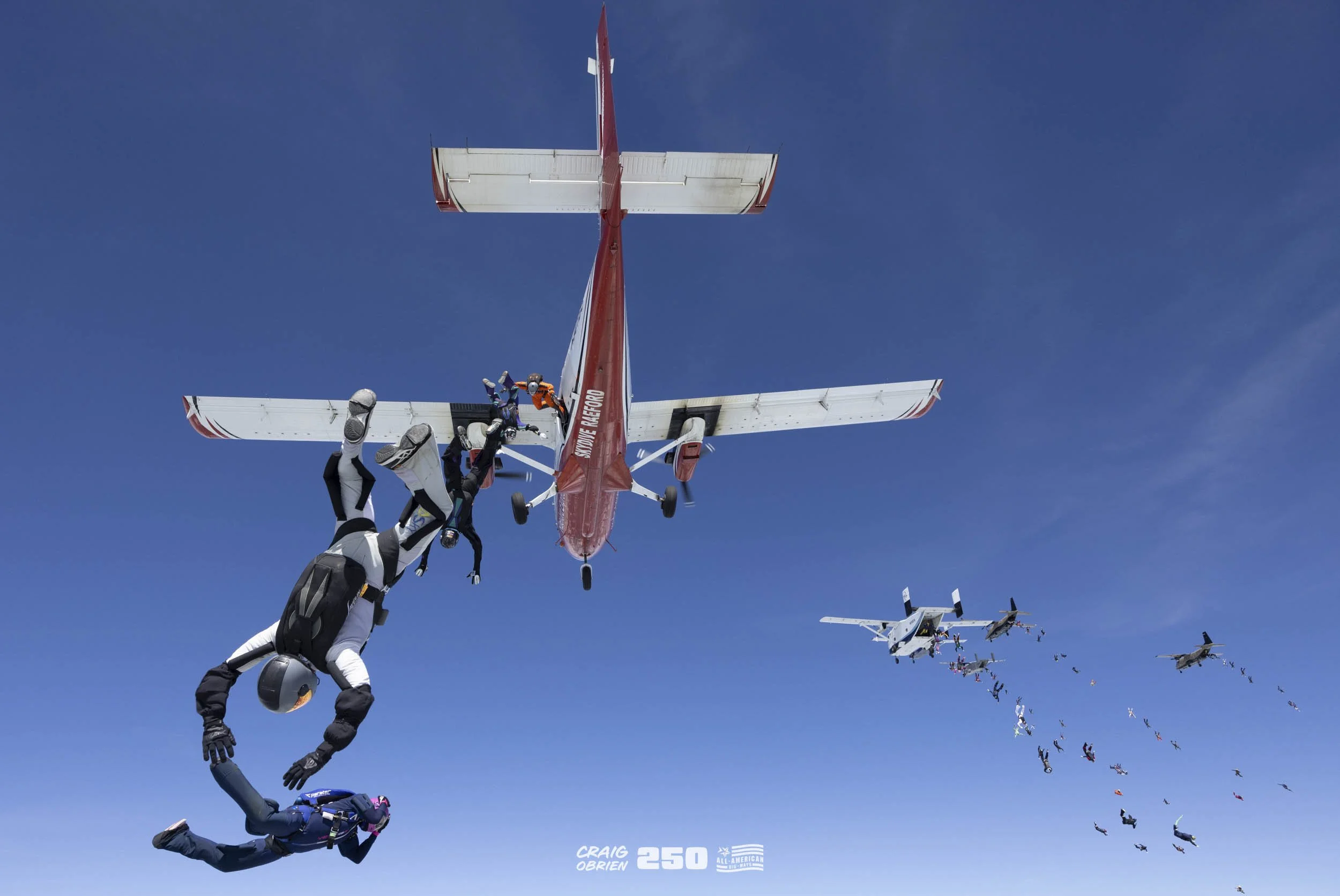 Skydivers in free fall formation with a red and white airplane in clear blue sky, parachutes open in background.