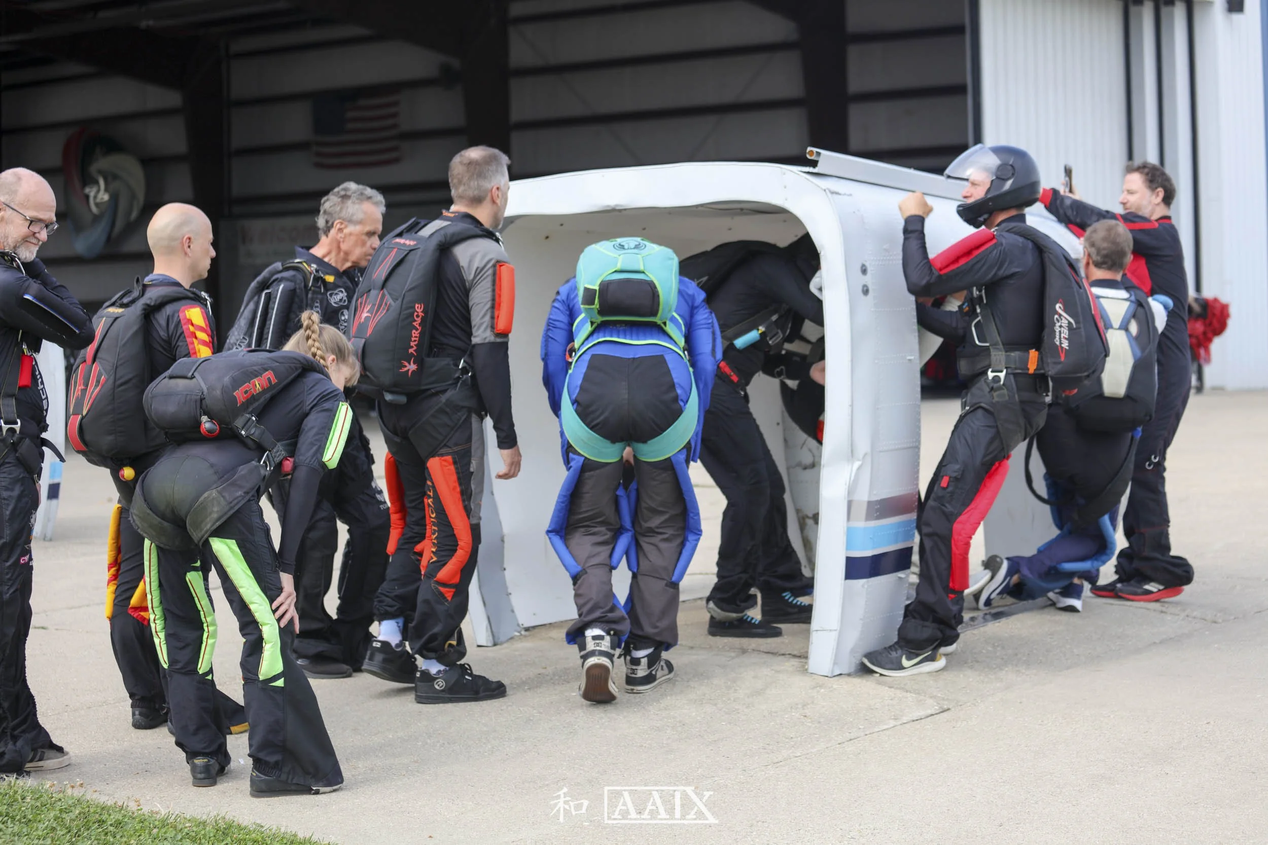 Skydivers preparing to board a small aircraft, with some entering the cockpit and others waiting in line with backpacks and jumpsuits.