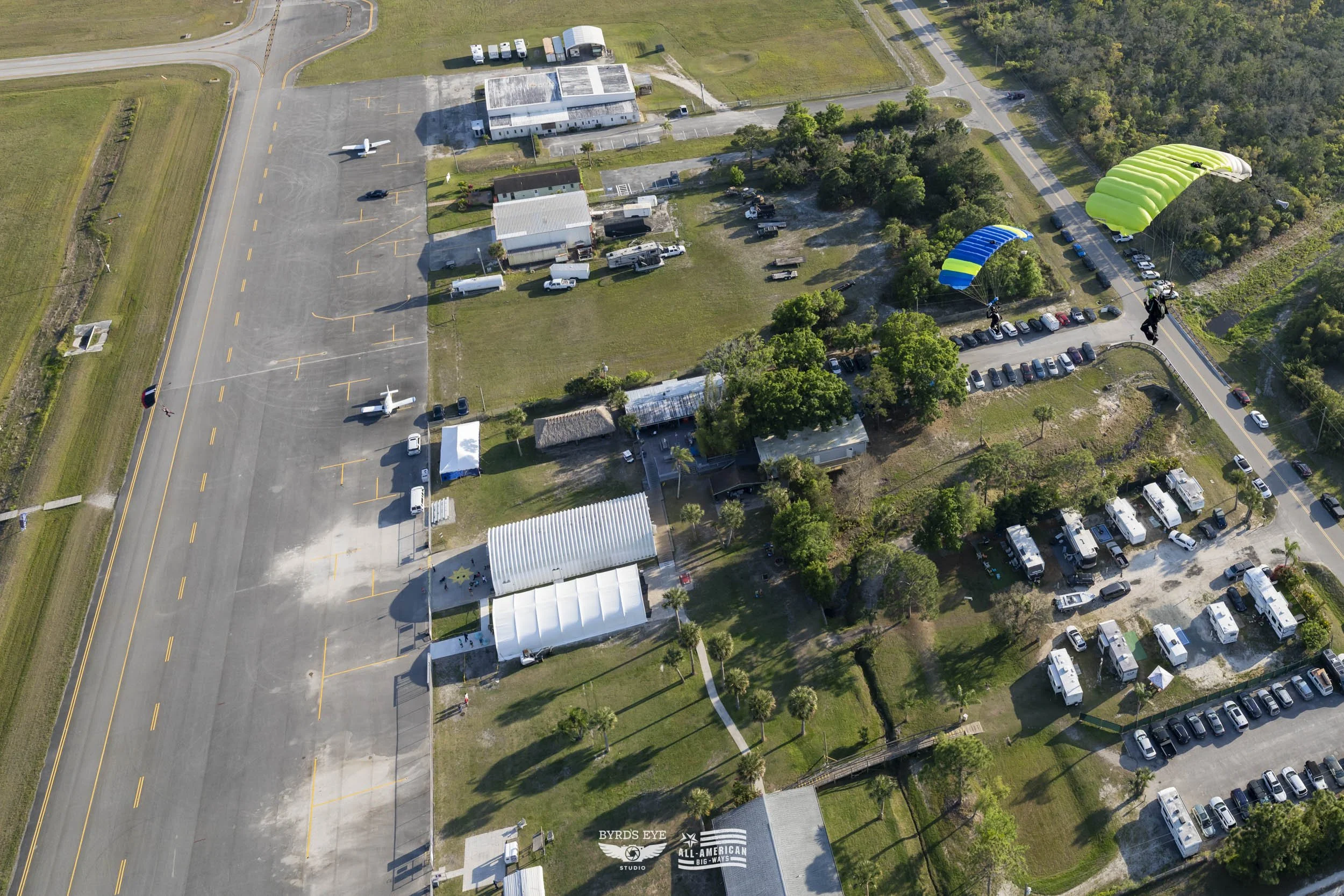 Aerial view of an outdoor area with parked aircraft, tents, vehicles, trees, and two people paragliding with bright green and blue parachutes.