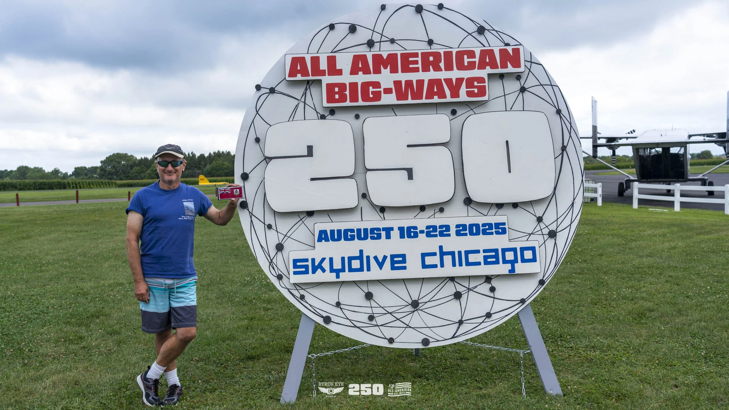 Man in blue shirt and shorts holding Canadian flag flag standing next to large sign at Skydive Chicago, celebrating 250th all-American big-ways jump from August 16-22, 2025.