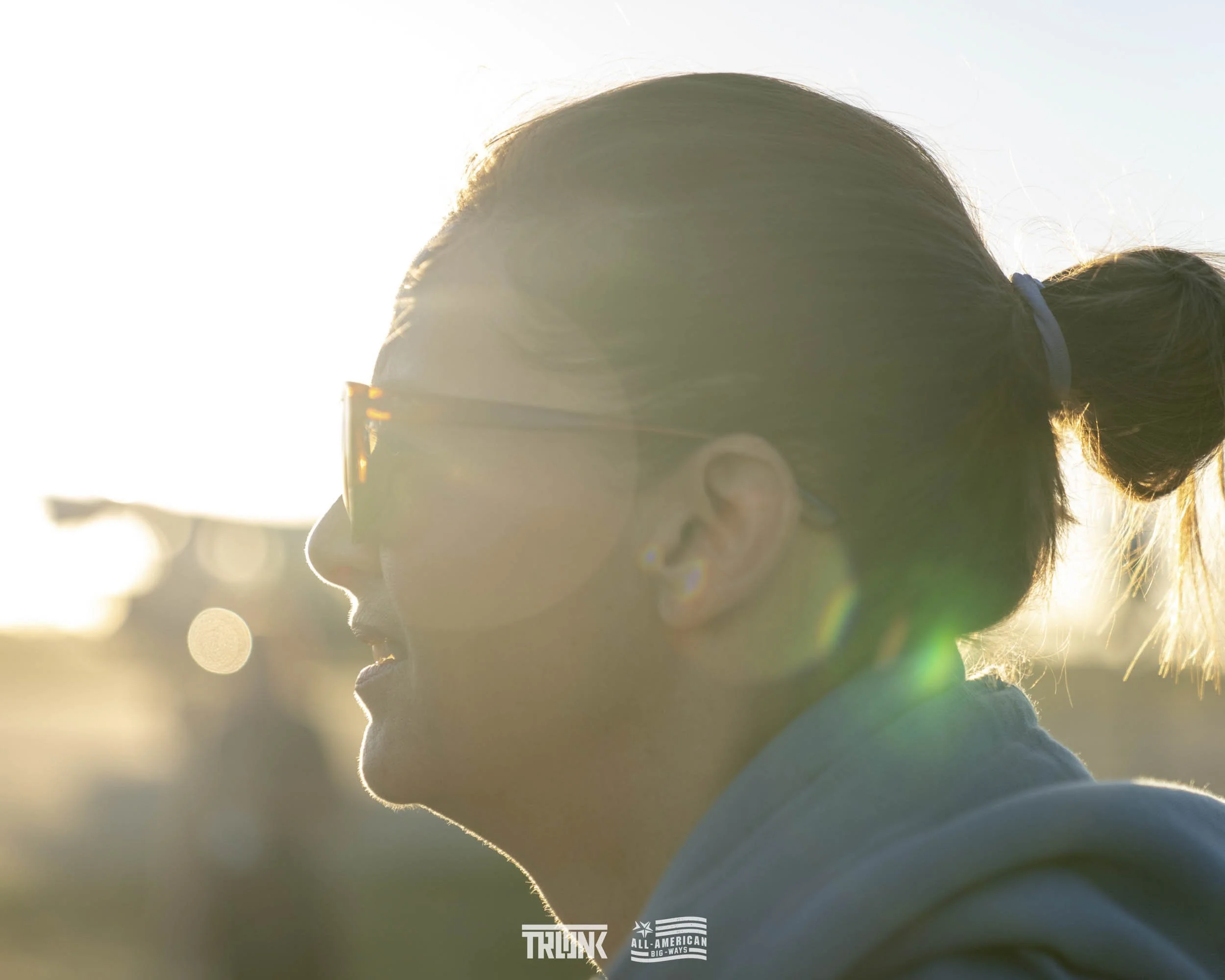 Profile of a young woman with glasses and a bun, backlit by the sun, outdoors in late afternoon or early evening.