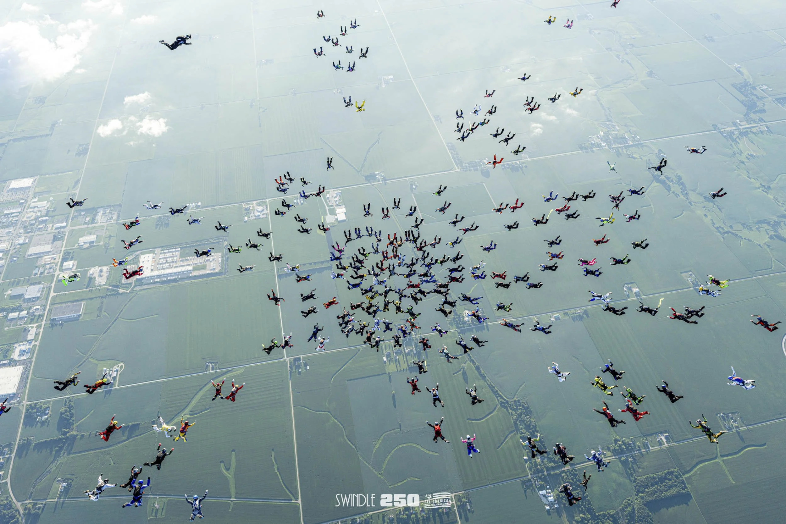 Aerial view of hundreds of skydivers forming a heart shape in the sky over a rural landscape. Some skydivers are in freefall, while others are descending with parachutes open. The landscape below includes fields, roads, and buildings, with a clear sk