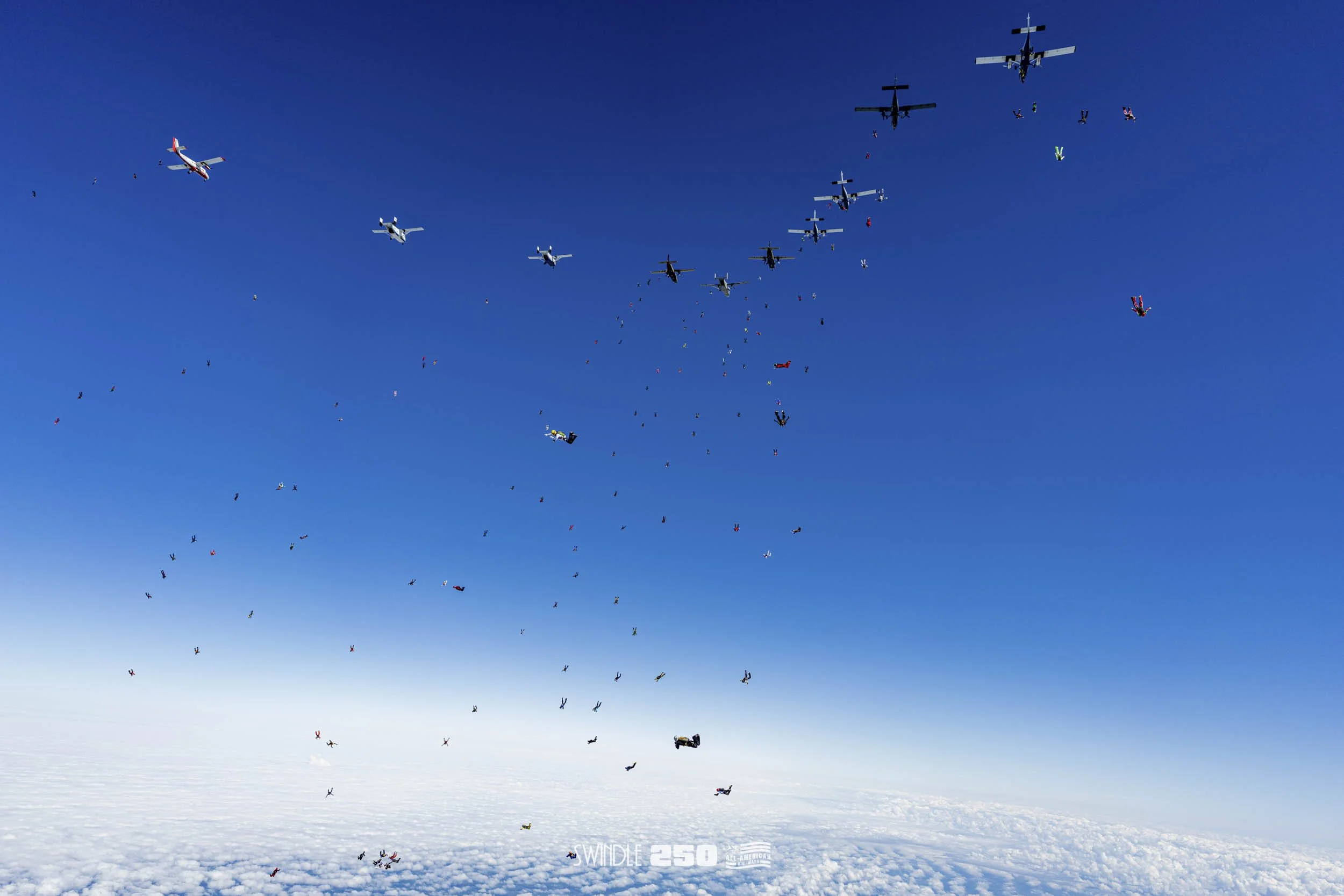 Skydivers free-falling from airplanes over a cloud-covered landscape, with planes flying above.