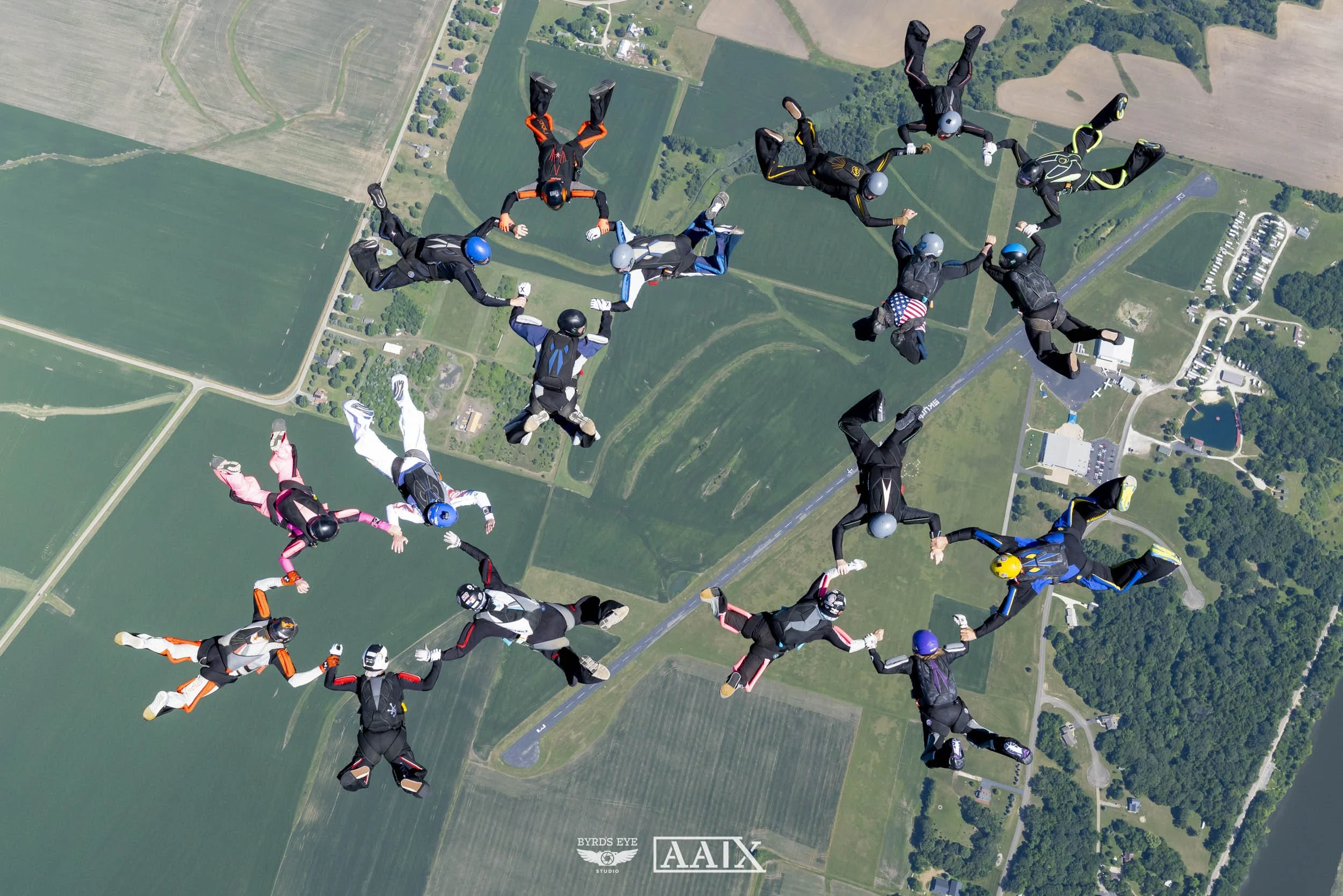 A group of skydivers holding hands forming a circle during free fall over a rural area with fields and roads.