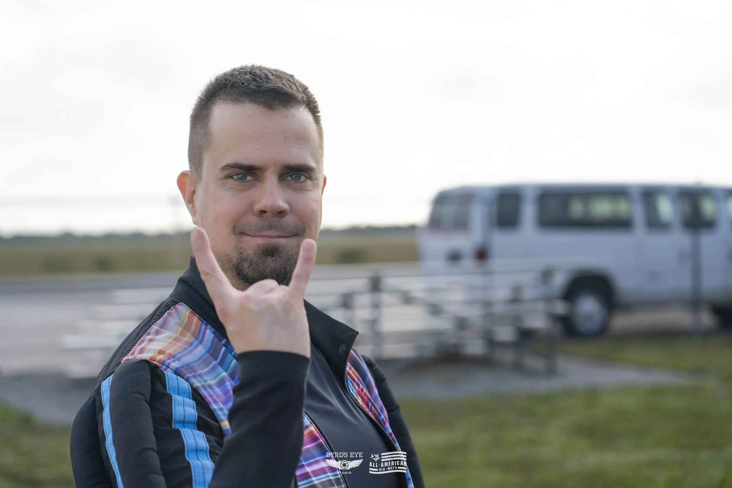 A man with short dark hair and a goatee, making the rock on hand gesture, standing outdoors near a vehicle, with a blurred background of a road and open field.