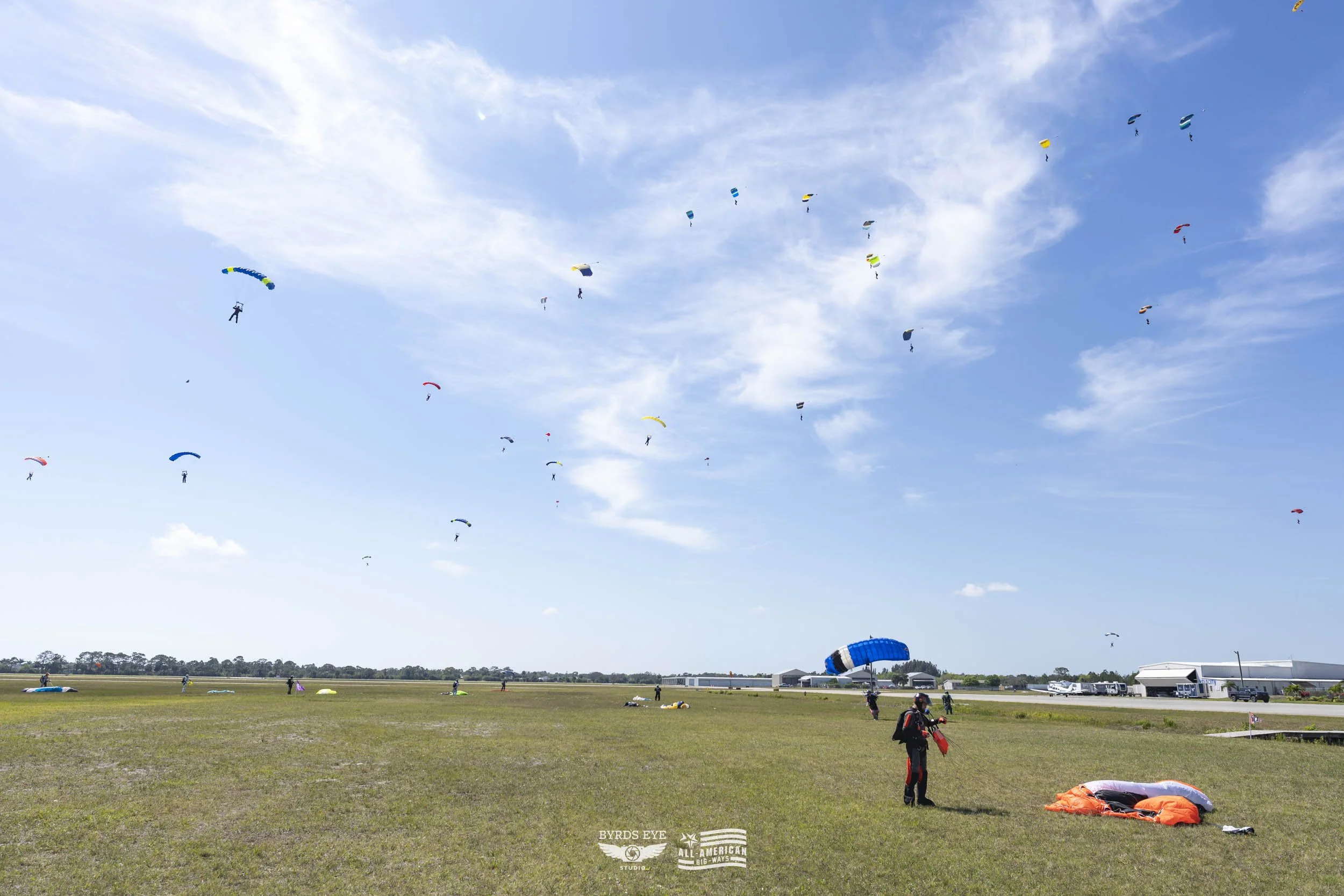 Several paragliders in colorful parachutes launching from a grassy field under a partly cloudy blue sky.