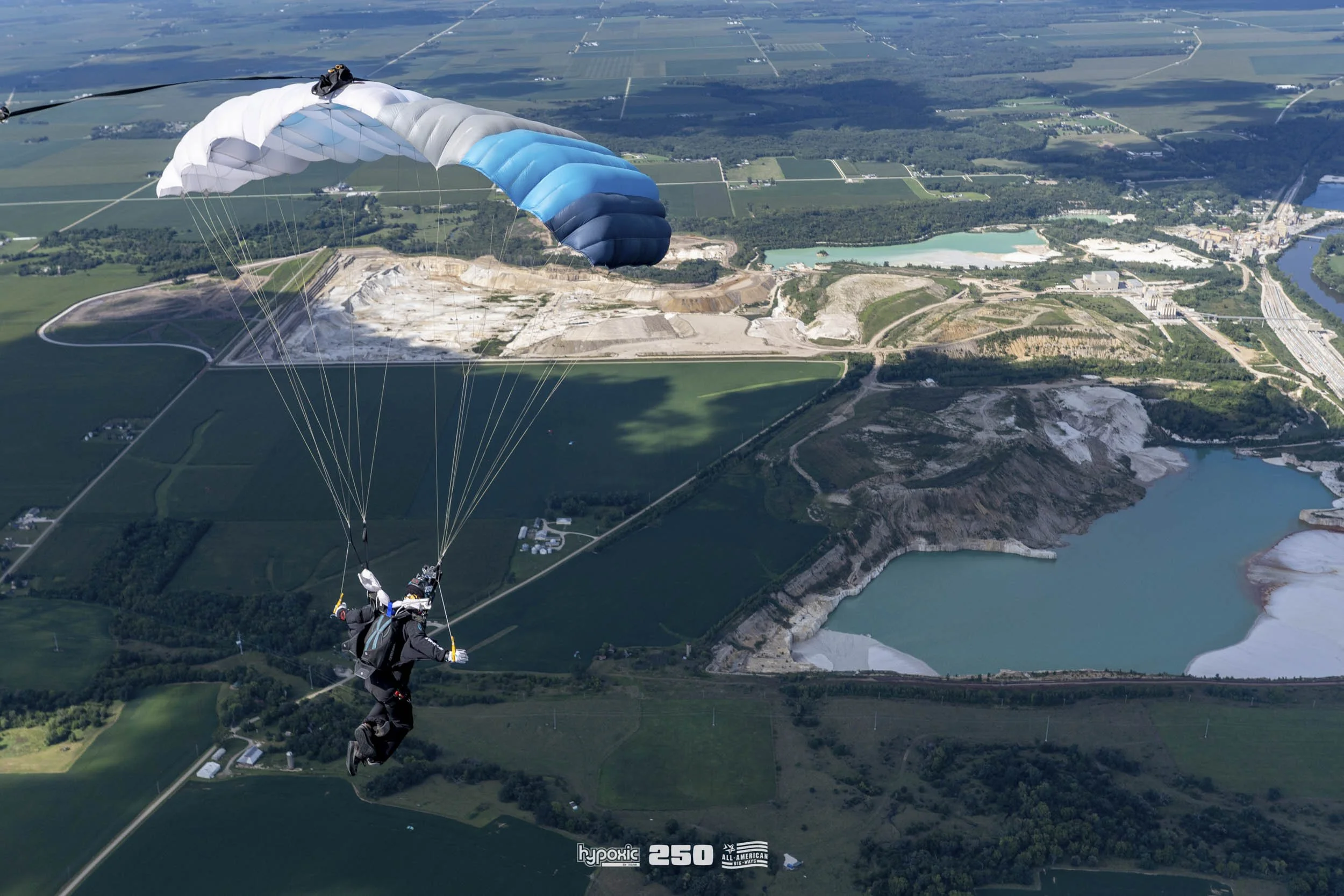 A person paragliding over a landscape with lakes, farmland, and a large open-pit quarry.
