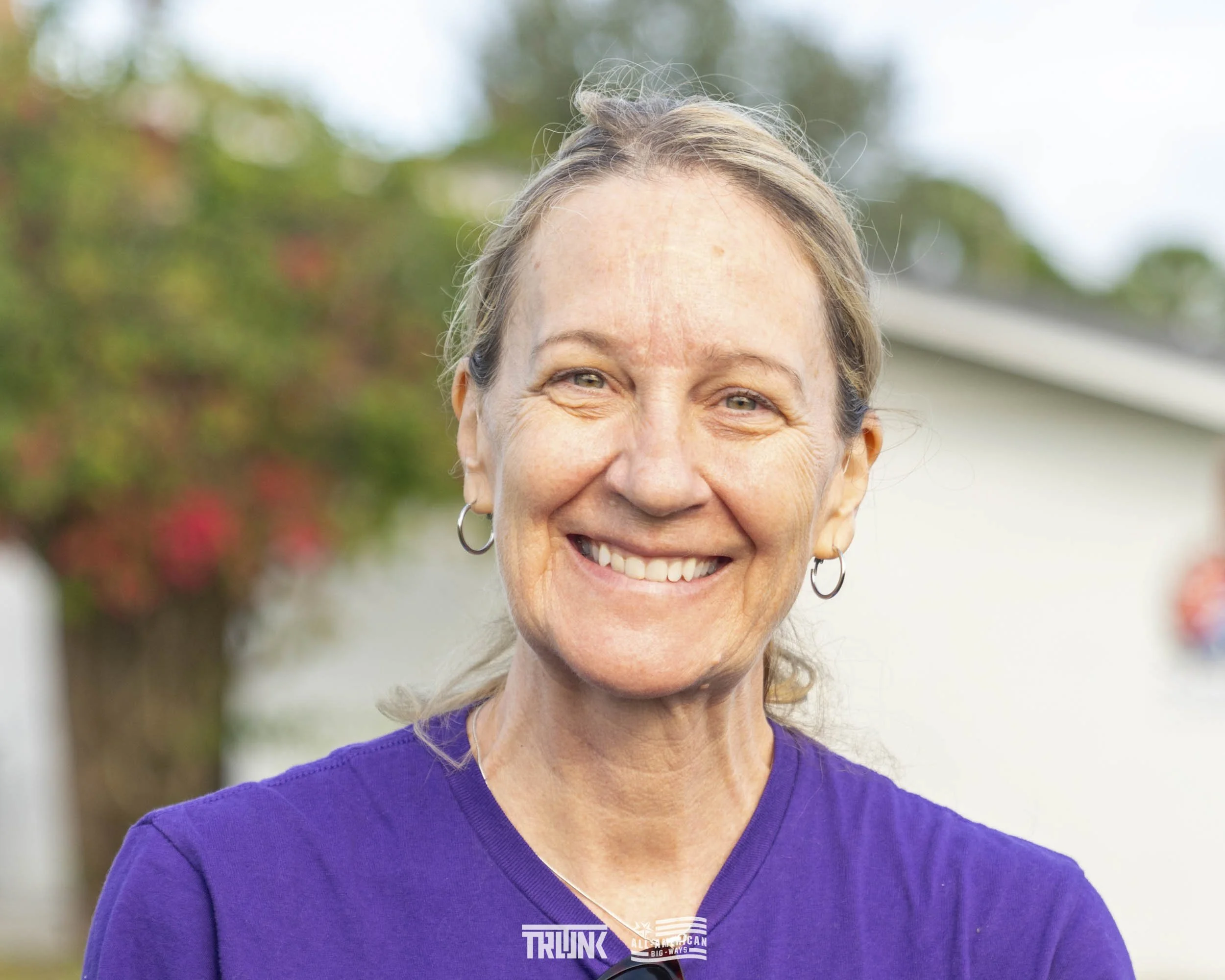 Smiling middle-aged woman with earrings wearing a purple shirt outdoors with trees in the background.