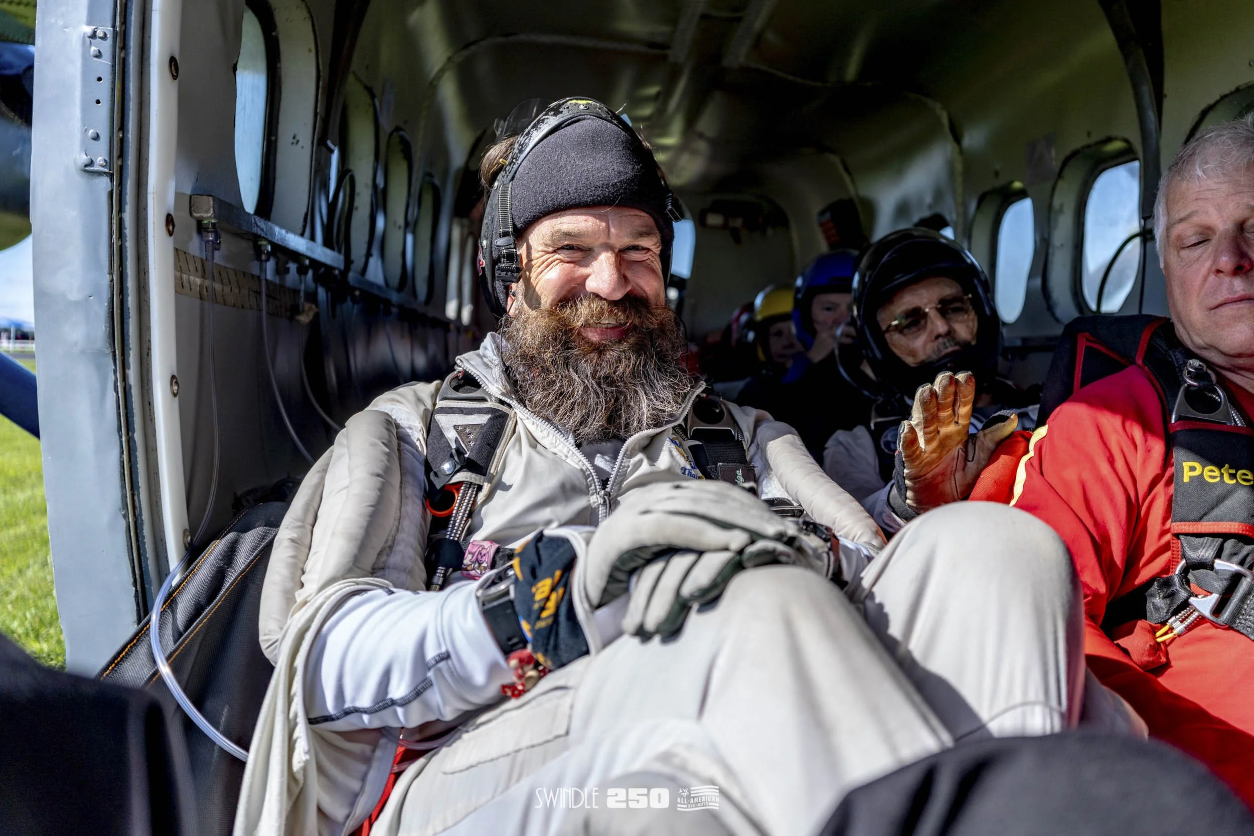 A group of skydivers sitting inside an aircraft, preparing for a jump. The man in the foreground is smiling and wearing a gray jumpsuit, gloves, and a helmet, with other skydivers in the background.