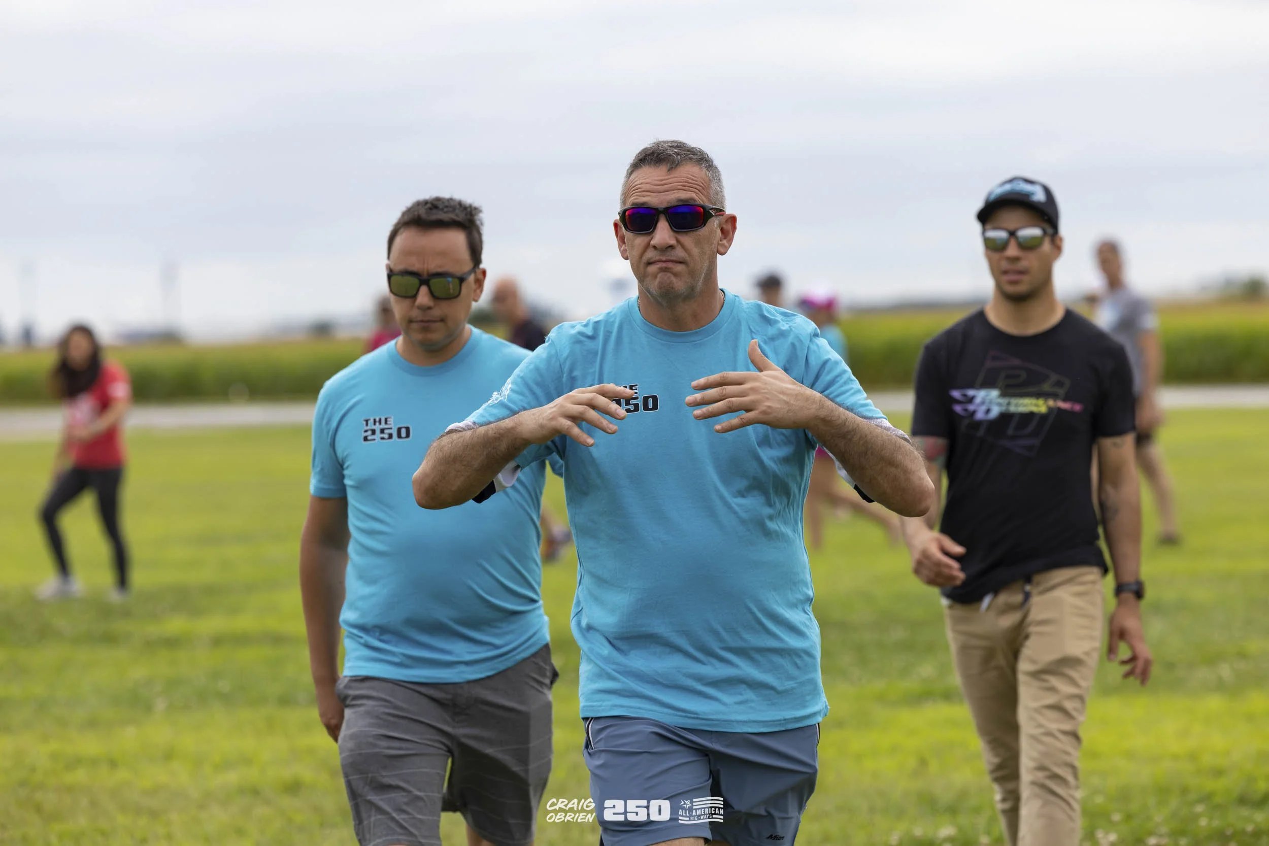 A group of people participating in an outdoor event, with three men in the foreground wearing sunglasses and T-shirts, and a woman in the background on a grassy field.