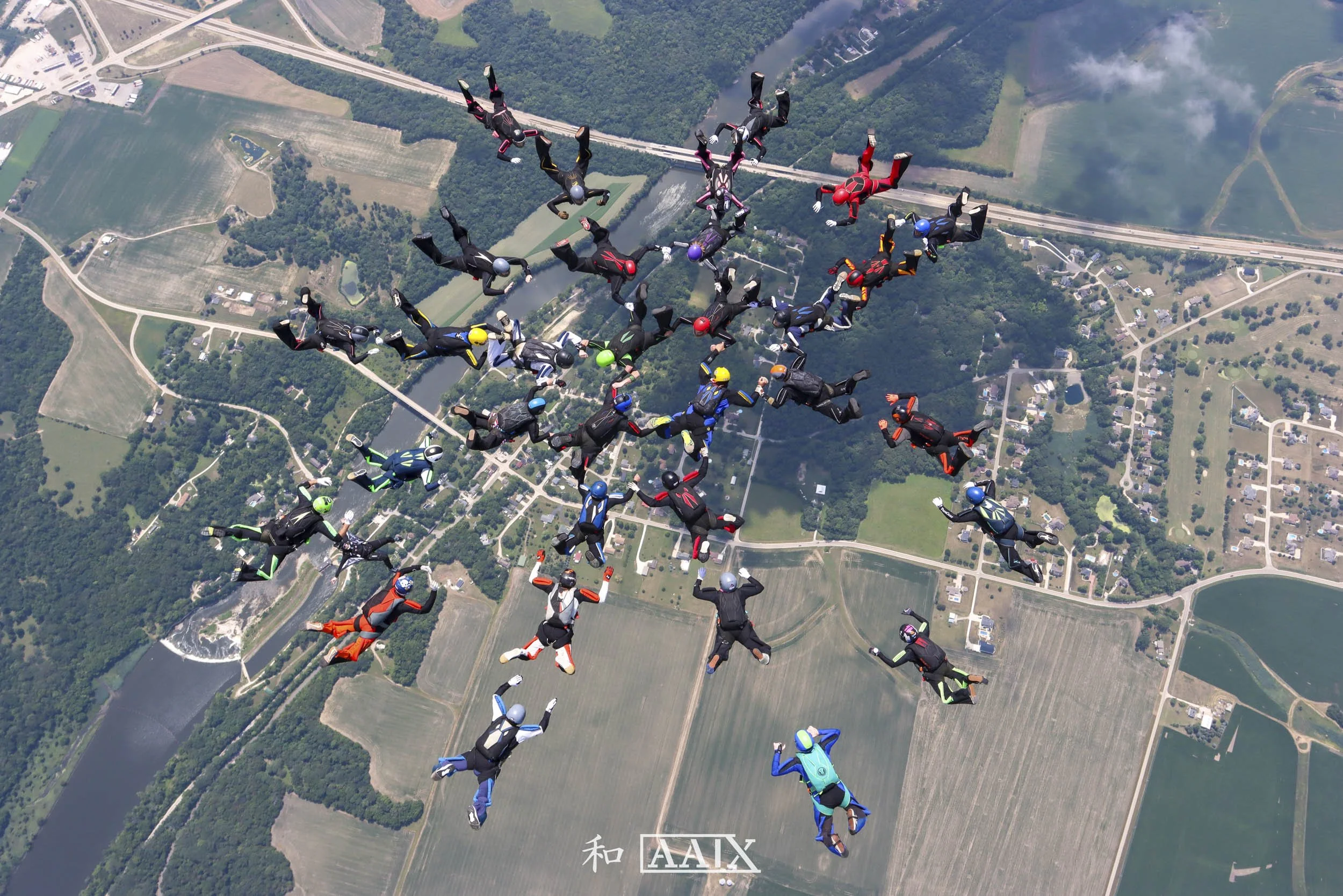 A large group of skydivers in colorful jumpsuits and helmets are free-falling in a formation above a patchwork of farmland, forests, and a river.