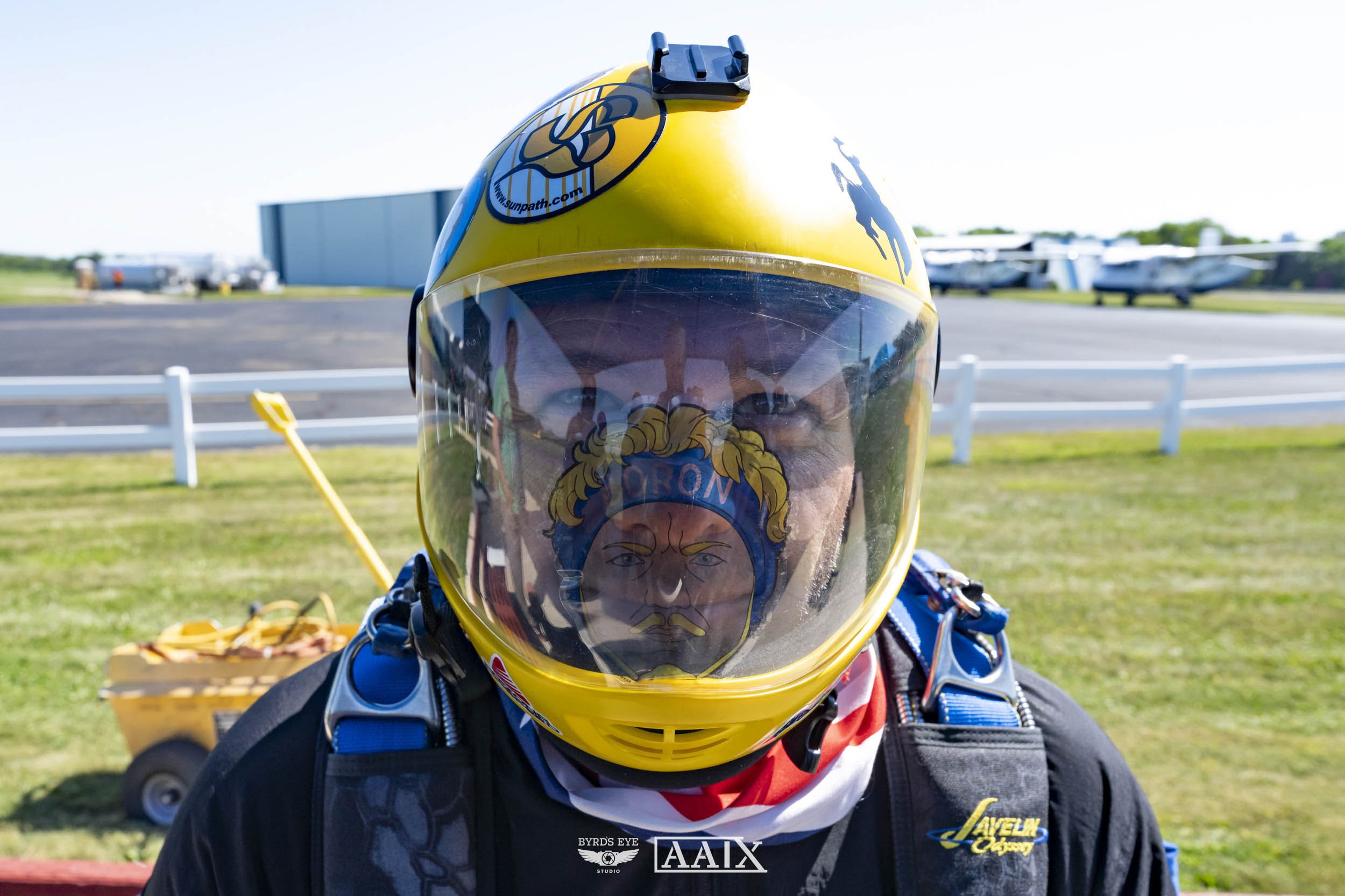 A person wearing a yellow racing helmet with a reflective visor, standing outdoors at an airfield with small planes in the background.