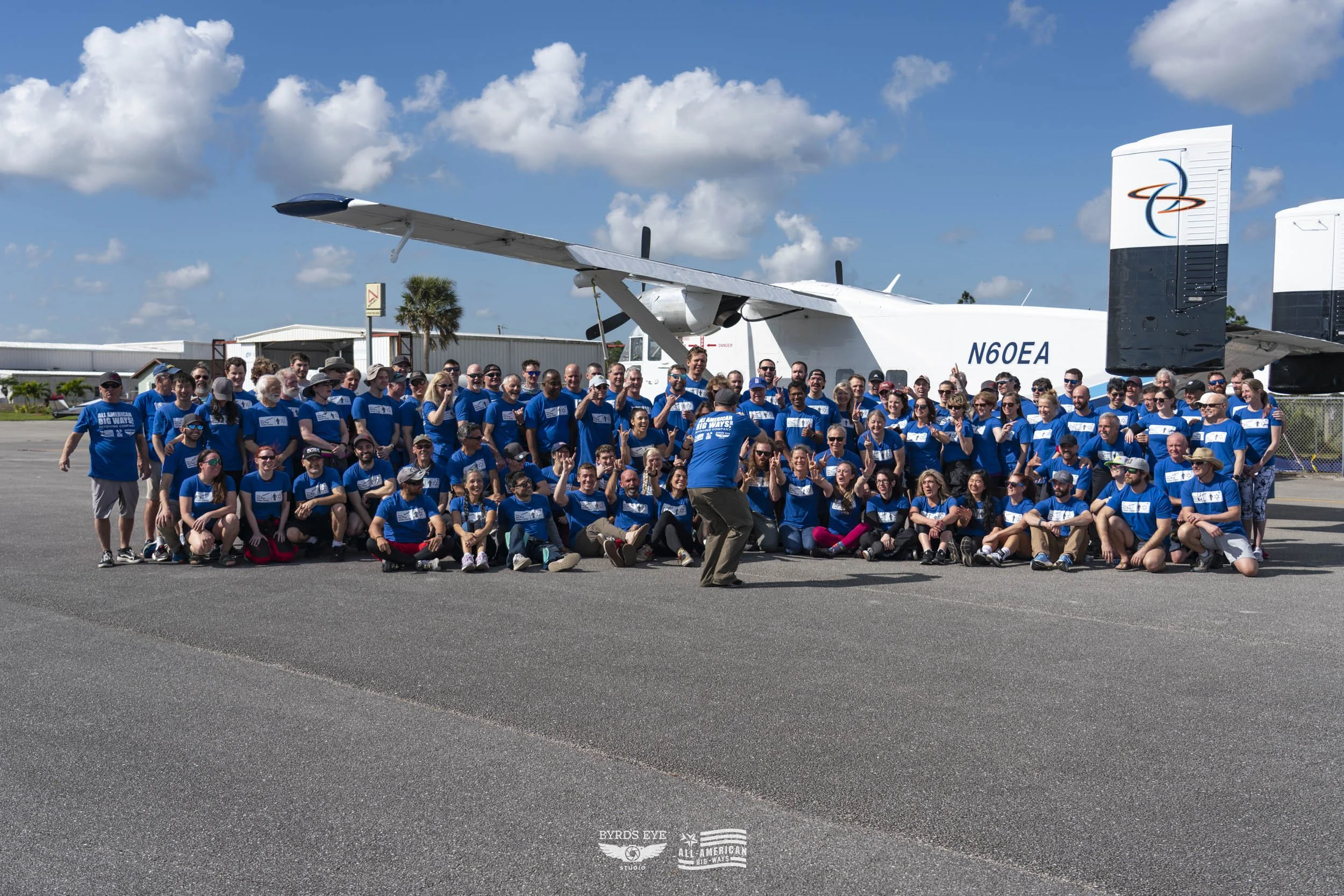 Group of people wearing blue shirts in front of a large white aircraft with the registration N60EA, on a sunny day with a partly cloudy sky.