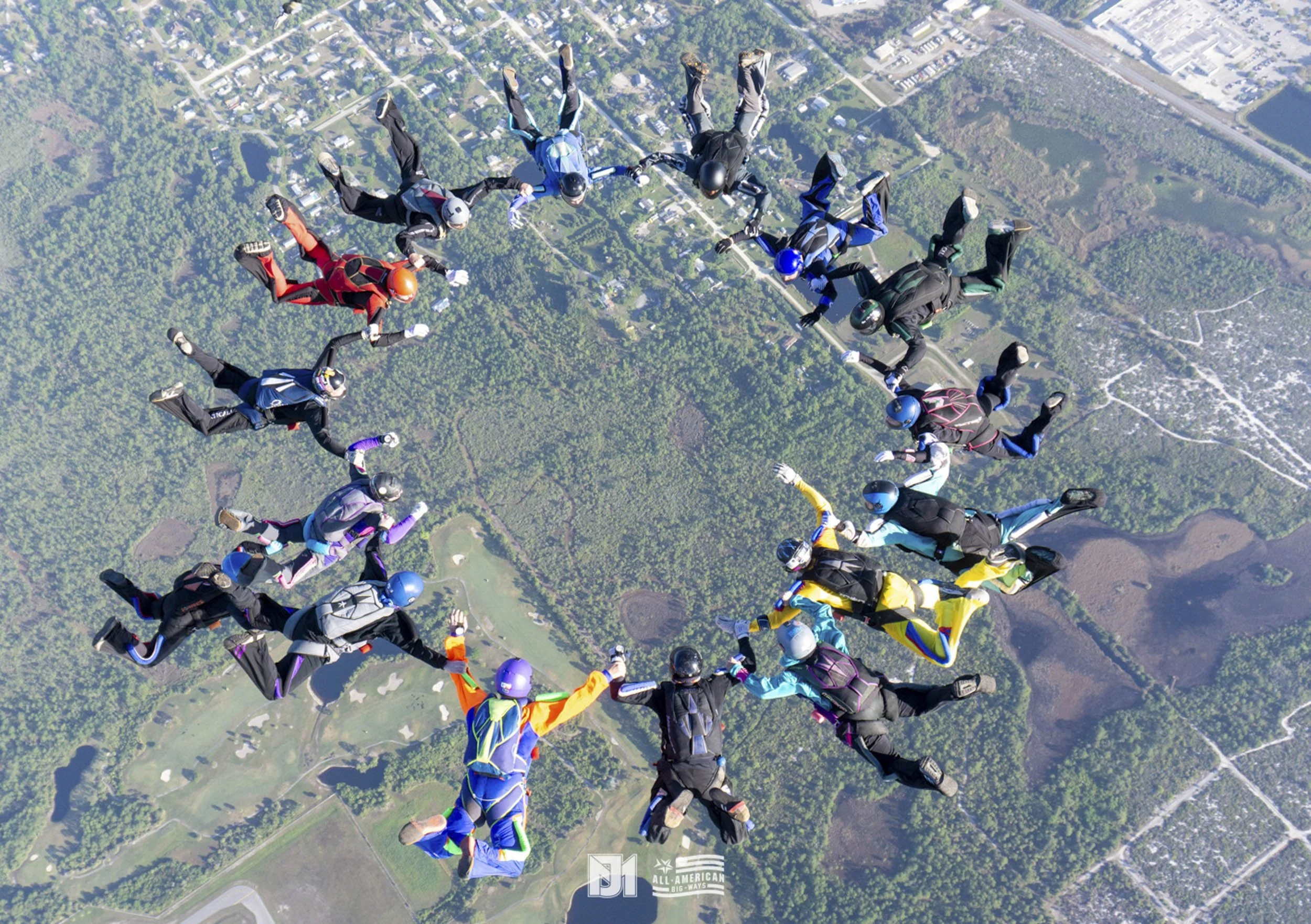 A group of skydivers forming a heart shape in mid-air over a landscape with trees, roads, and bodies of water.