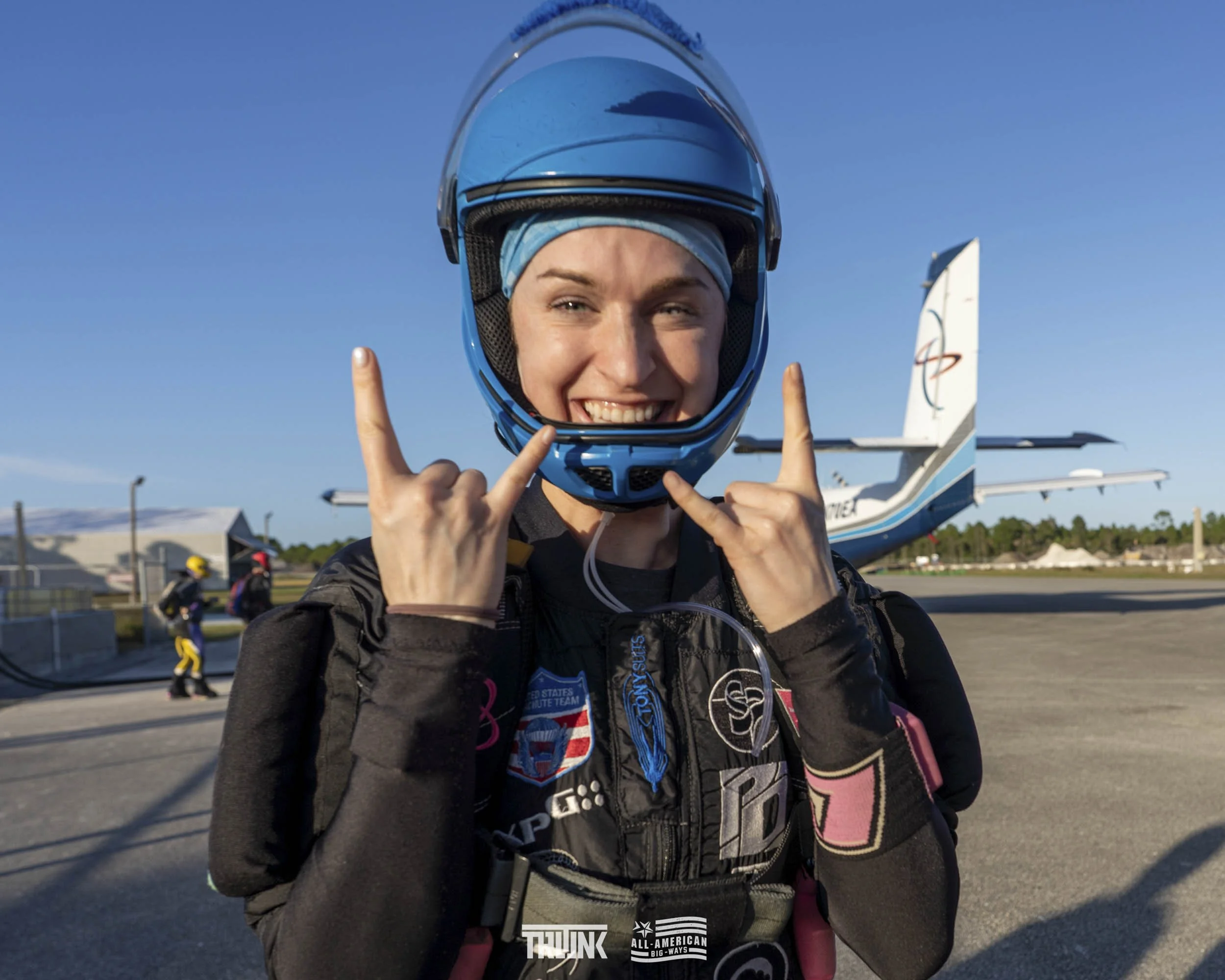 A smiling woman in skydiving gear, wearing a helmet, making the 'rock on' hand gesture, standing on the tarmac with an airplane in the background.