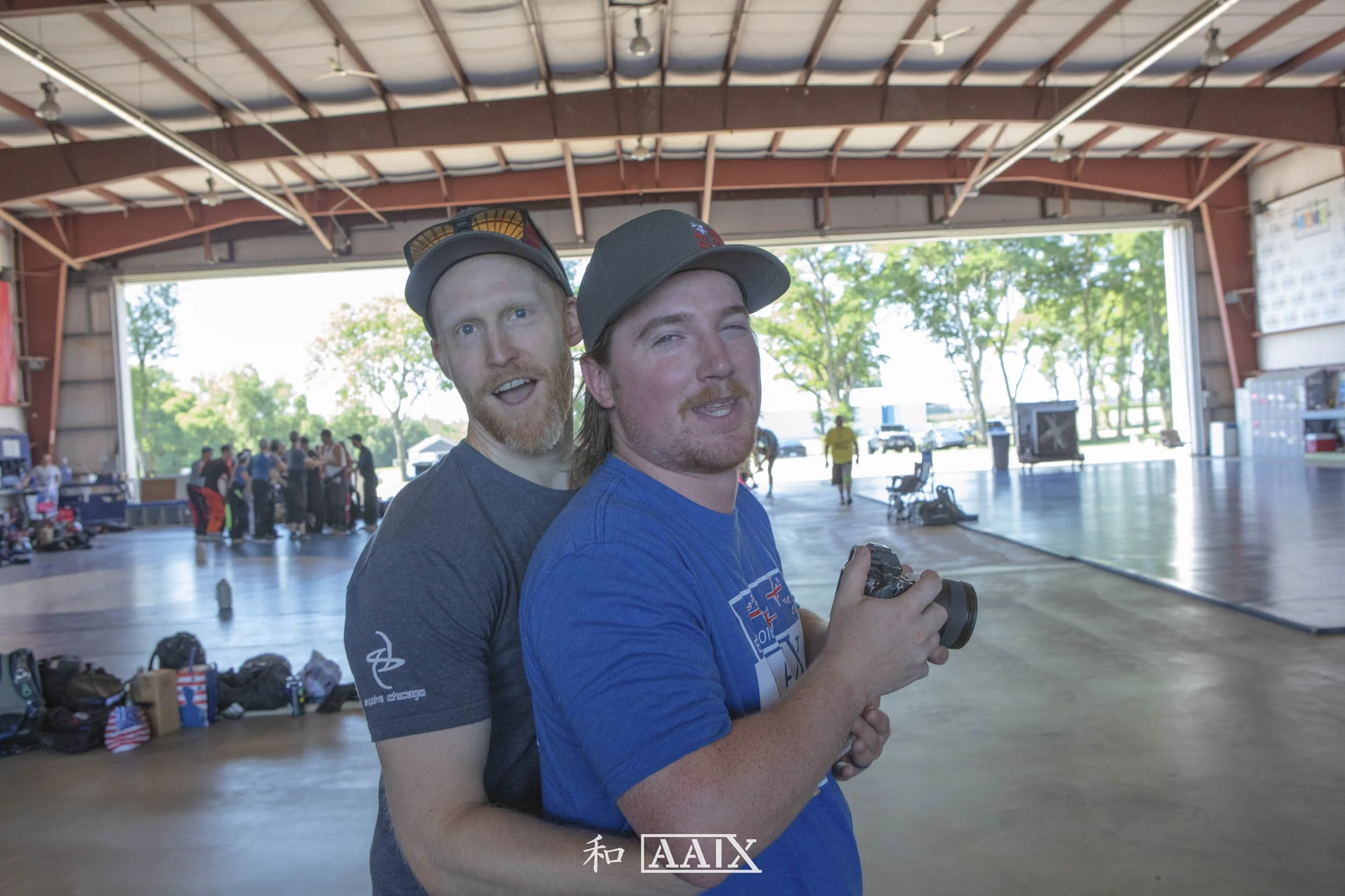 Two young men with casual clothing and baseball caps, one holding a camera, standing inside a large open hangar or gymnasium with other people and equipment in the background.