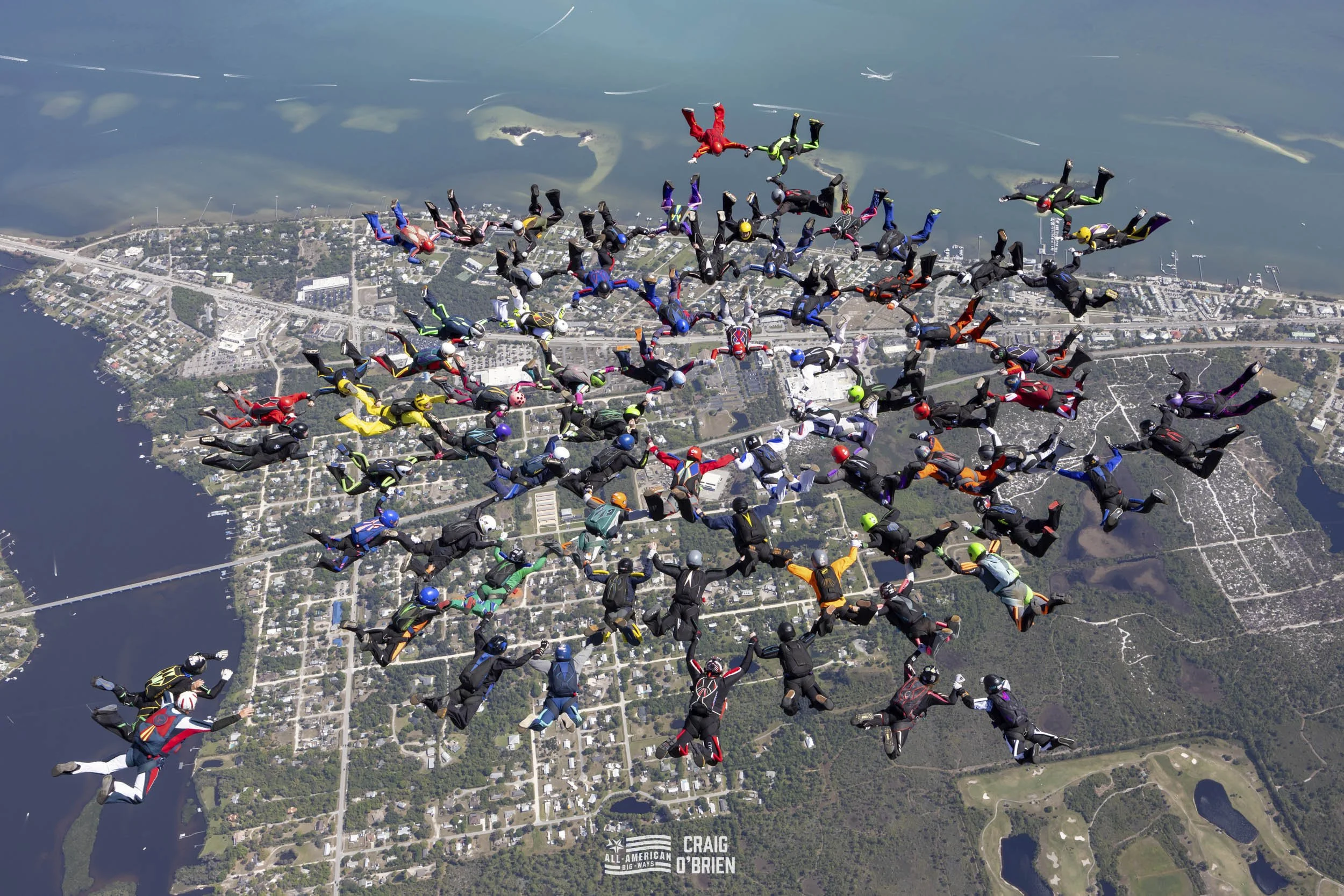 Group of skydivers in colorful jumpsuits skydiving over a landscape with lakes, roads, and trees.