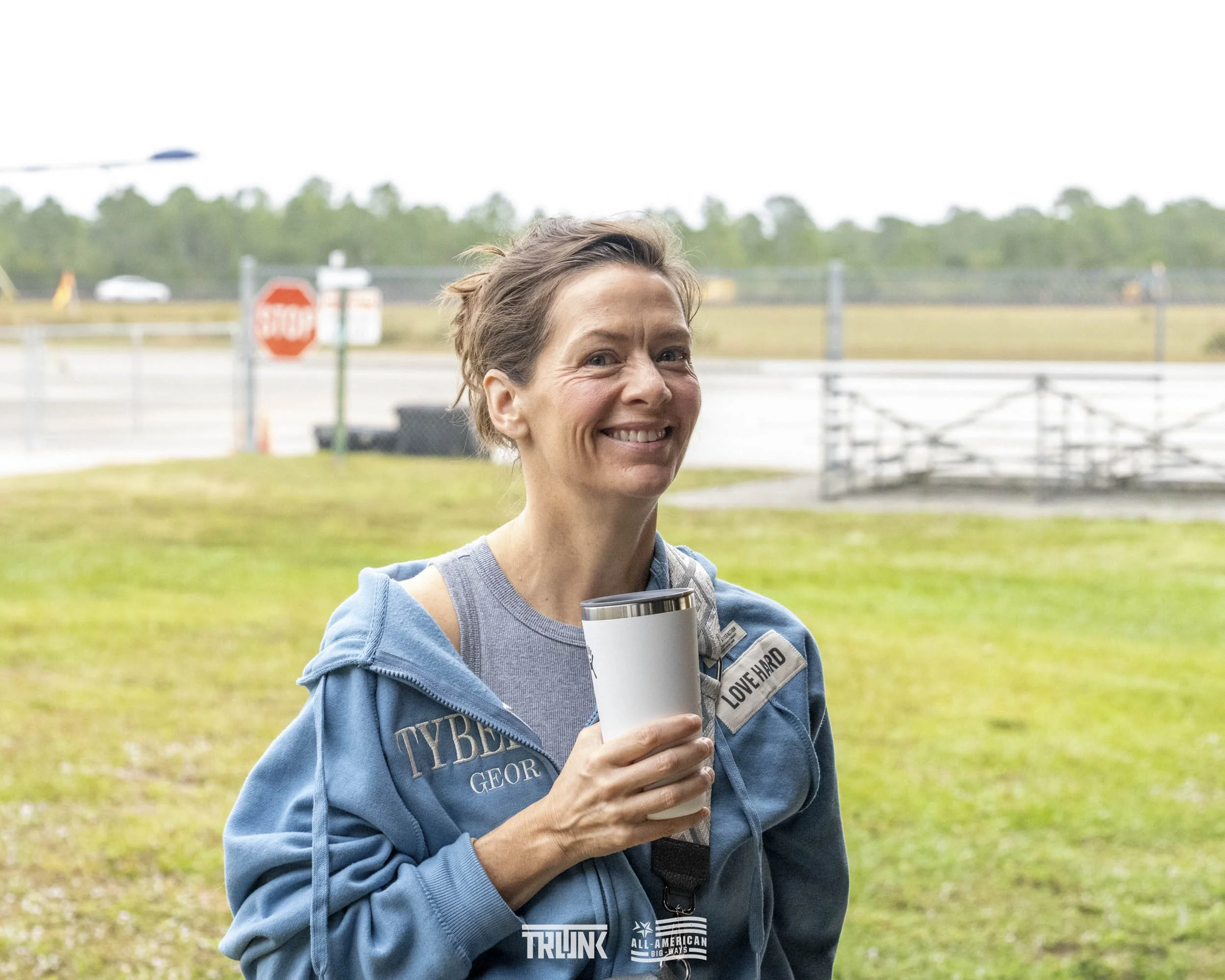 A smiling woman standing outdoors holding a stainless steel tumbler, wearing a blue jacket with name tags, on a cloudy day with a grassy area and a fenced waterway in the background.