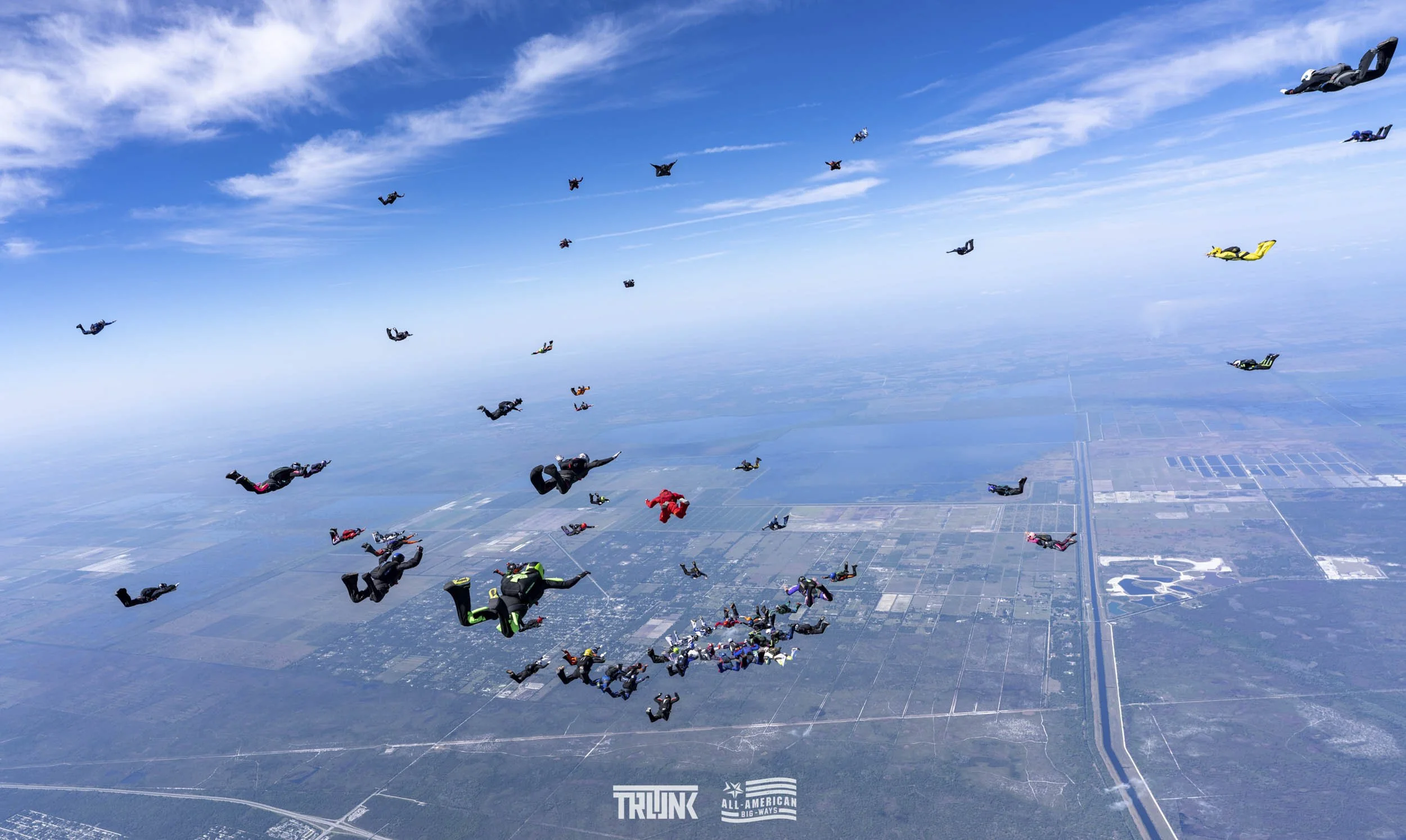 A group of skydivers in free fall over a landscape with fields and a highway, with some skydivers closer to the camera and others farther away, under a blue sky with scattered clouds.