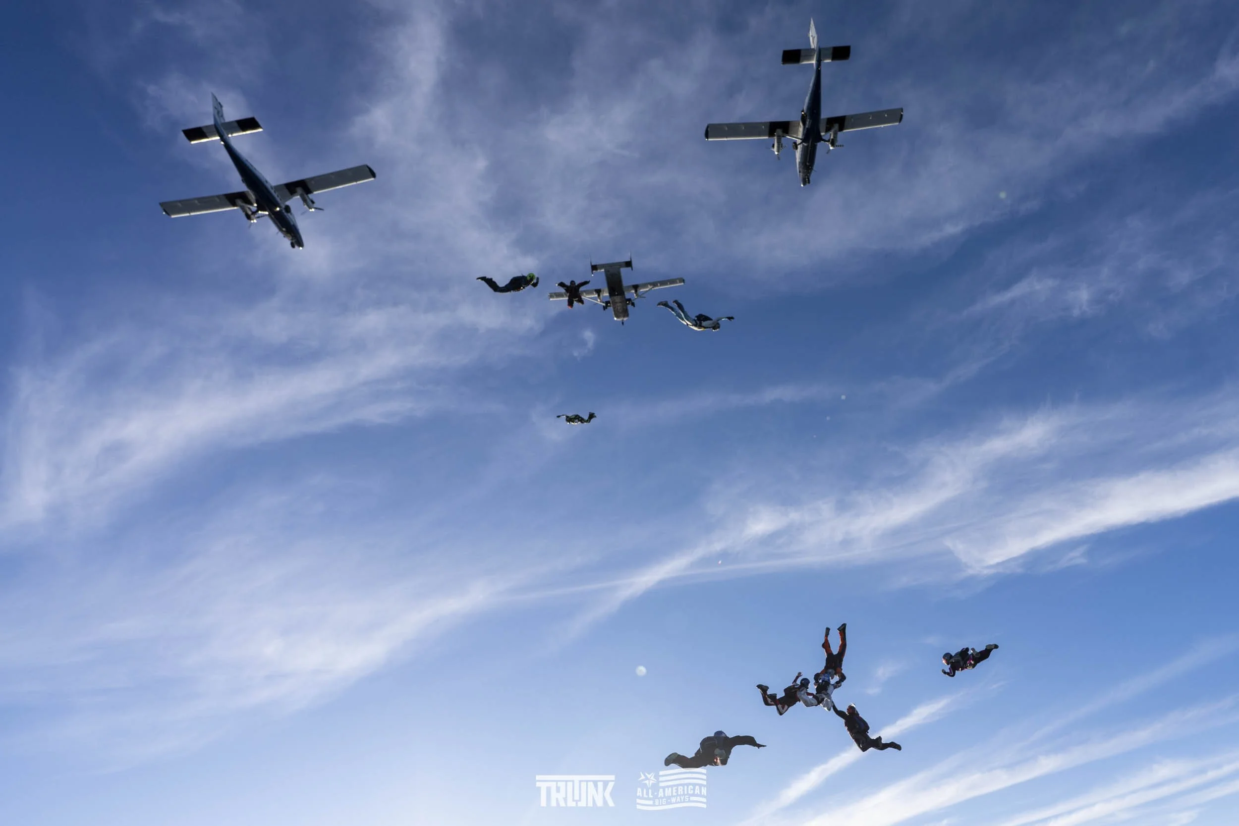 Skydivers in free fall with some airplanes flying overhead in a blue sky with wispy clouds.