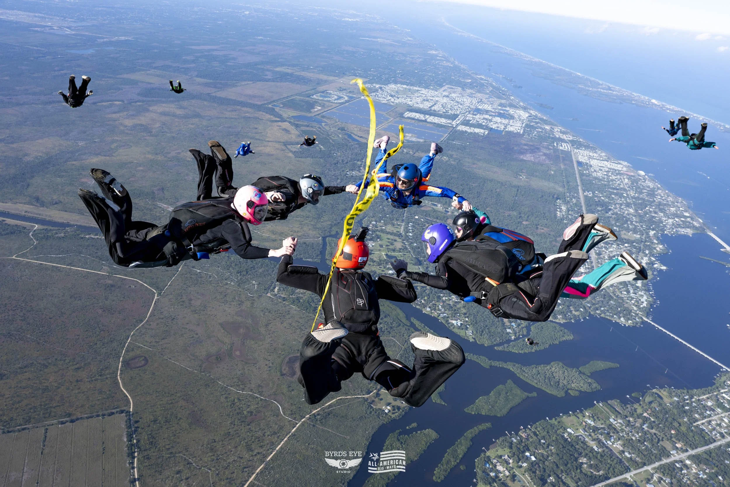 Group of skydivers holding hands in freefall over a landscape of water, forests, and urban areas with a coastline in the background.