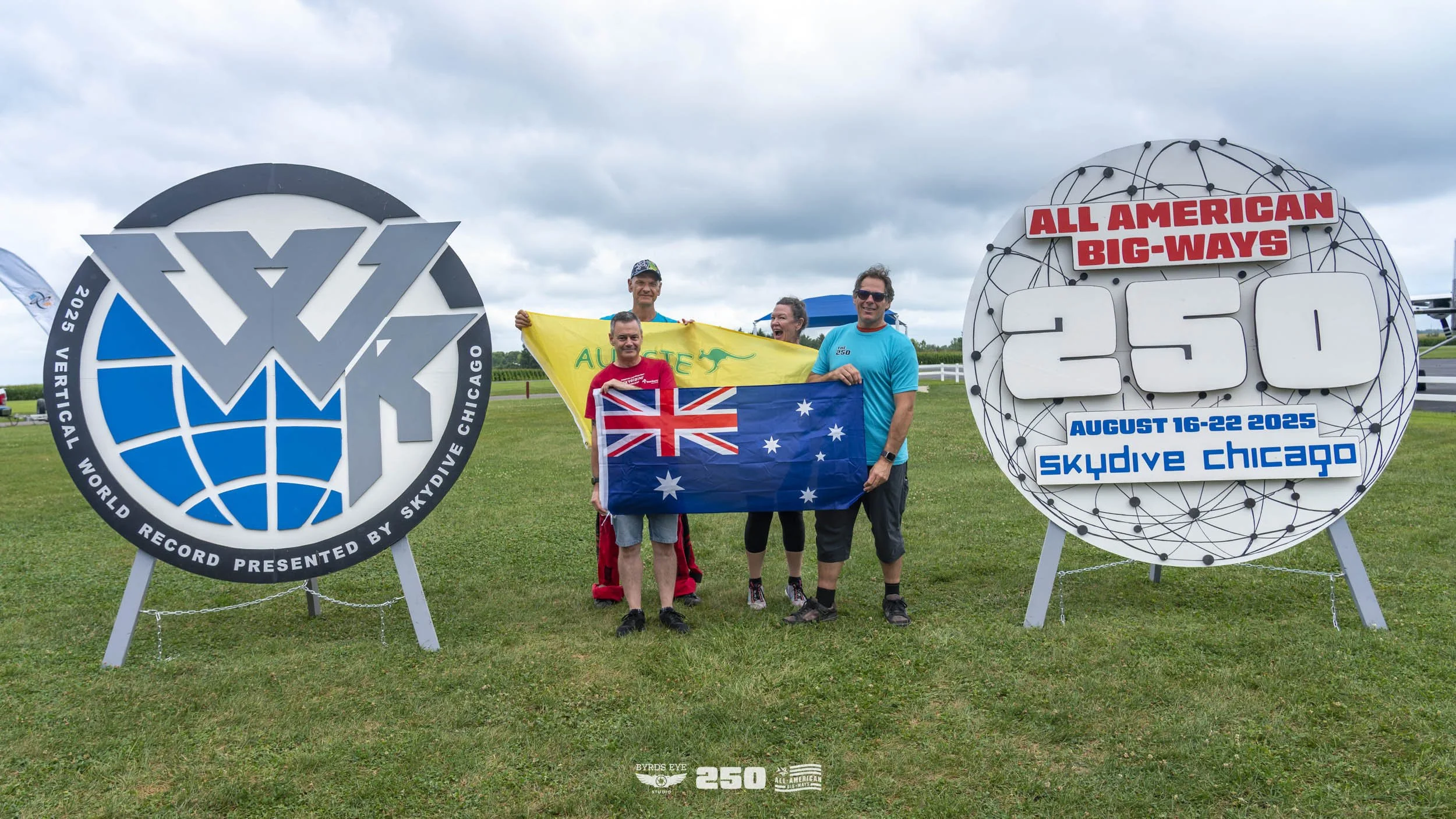 Group of five people standing on grass holding Australian and British flags, celebrating near two large signs for the All American Big-Ways event. One sign shows a world record for skydiving with a logo, the other displays event details including dat