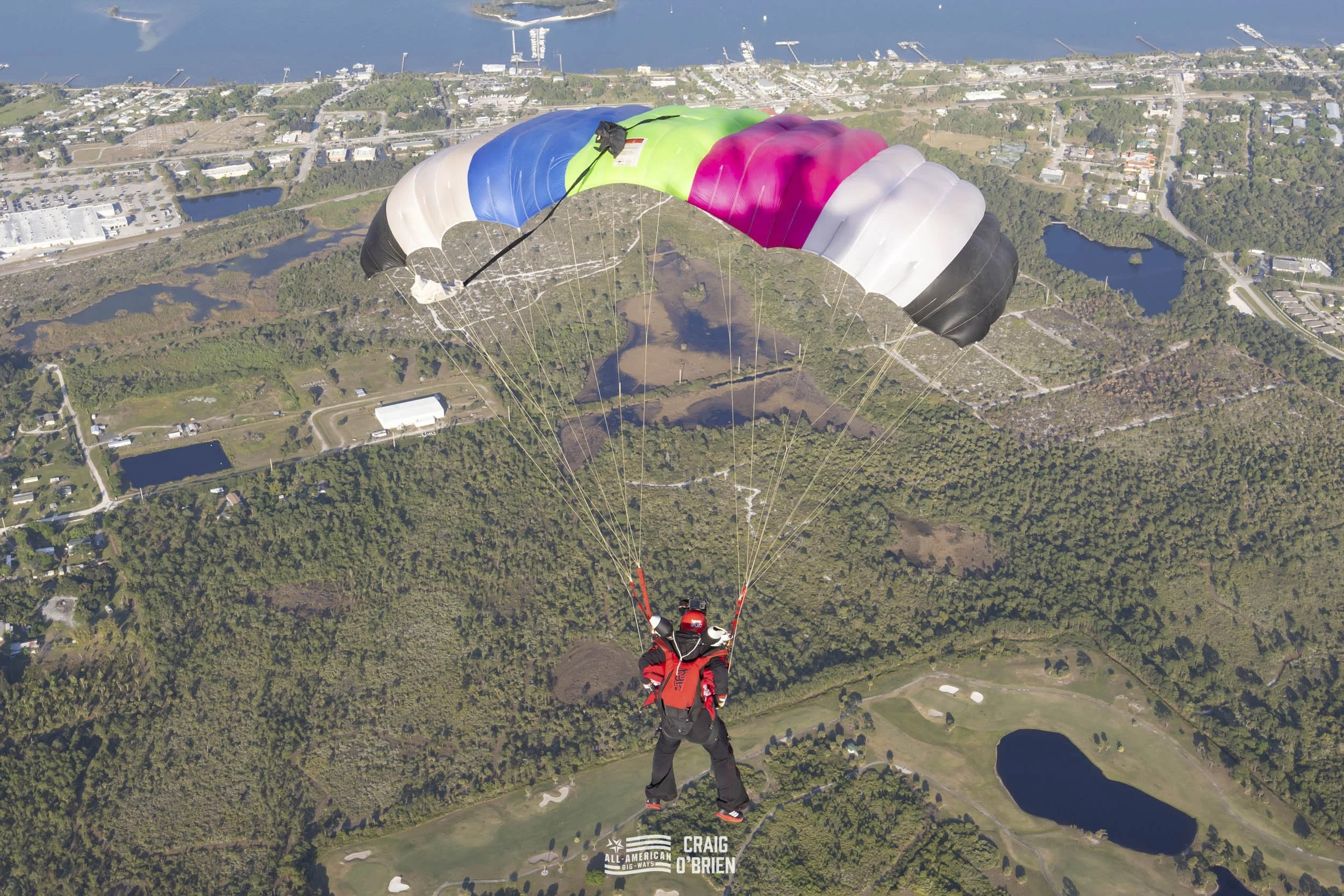A skydiver with a red and black suit and helmet descending with a colorful parachute over a landscape of green fields, lakes, and residential areas.