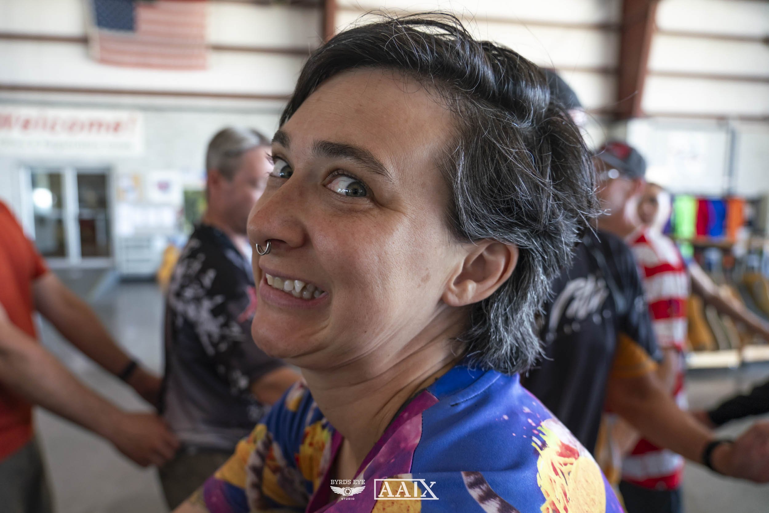 A woman with short, grayish hair, wearing a colorful shirt and a septum piercing, smiling and looking to the side in a busy indoor setting with other people in the background.
