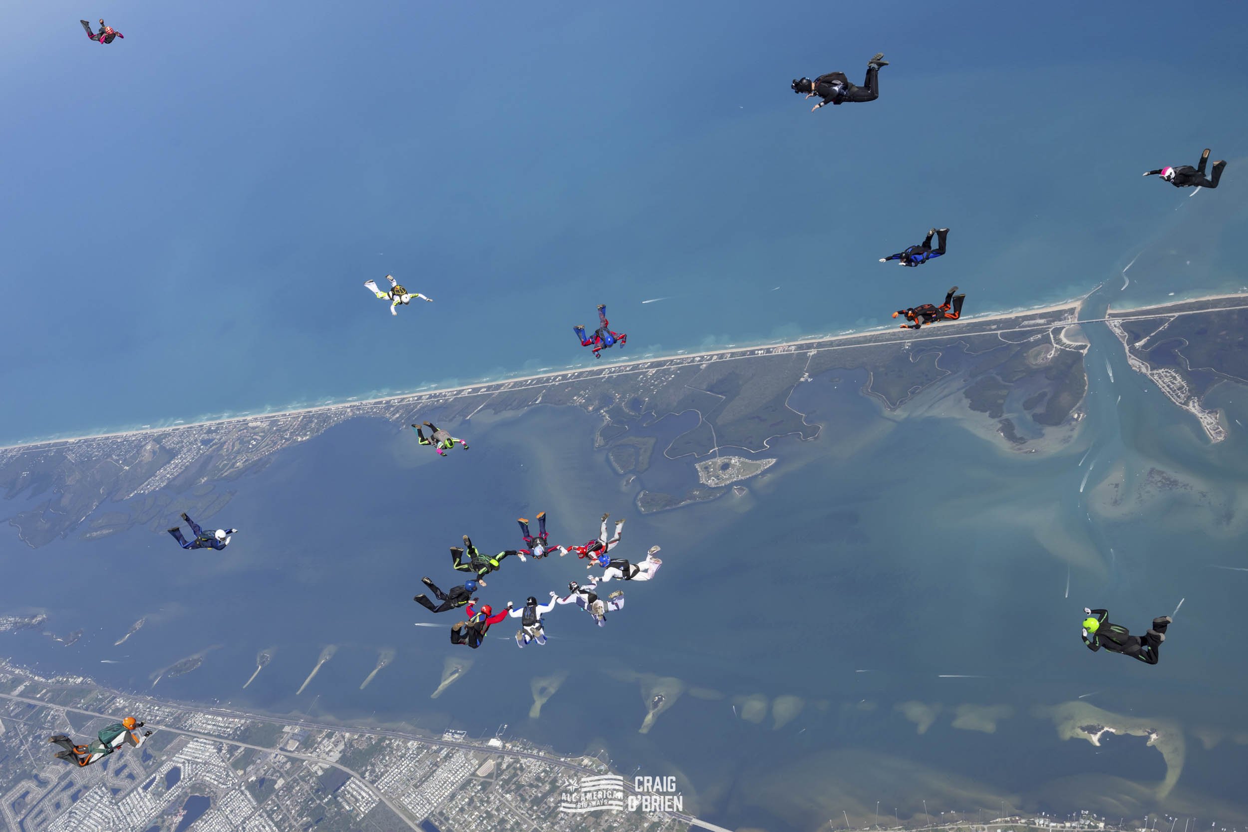 Skydivers in free fall above a coastal landscape, forming a circle and holding hands mid-air.