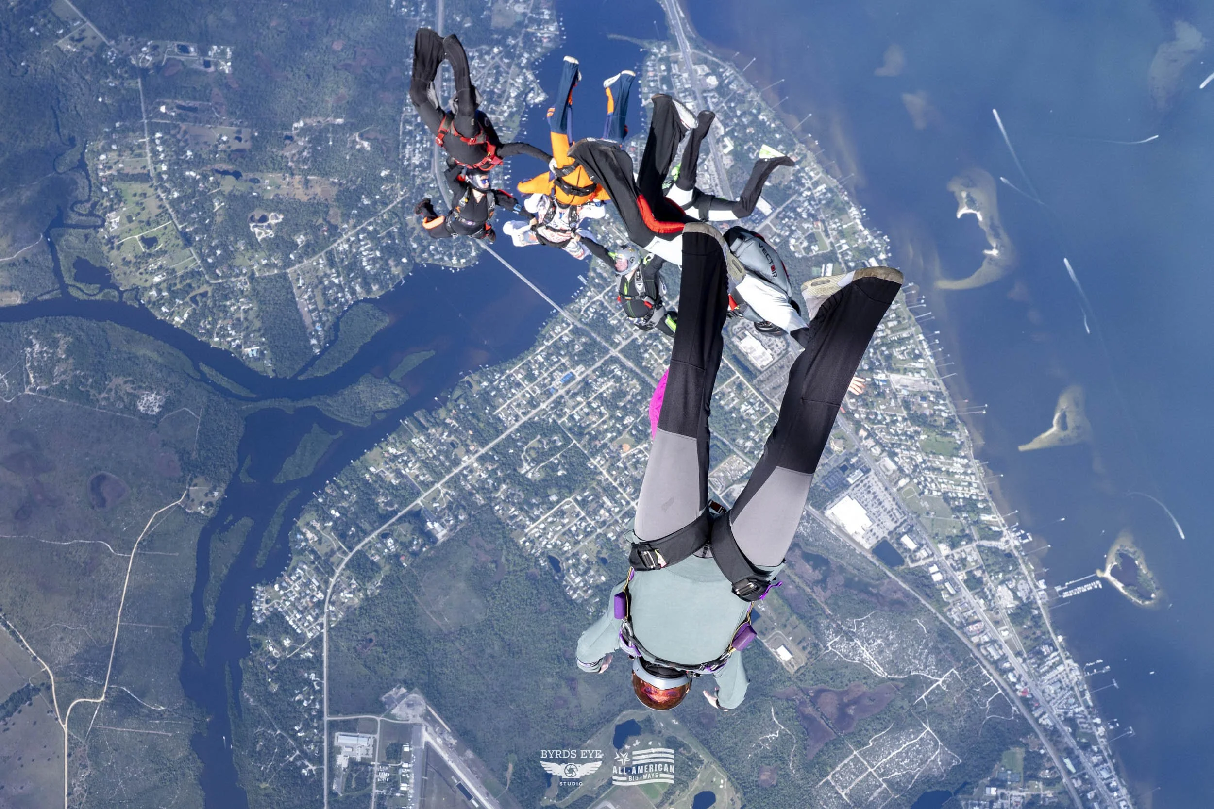 Skydiver free-falling over a city with a river and harbor, wearing a helmet and jumpsuit, surrounded by other skydivers.