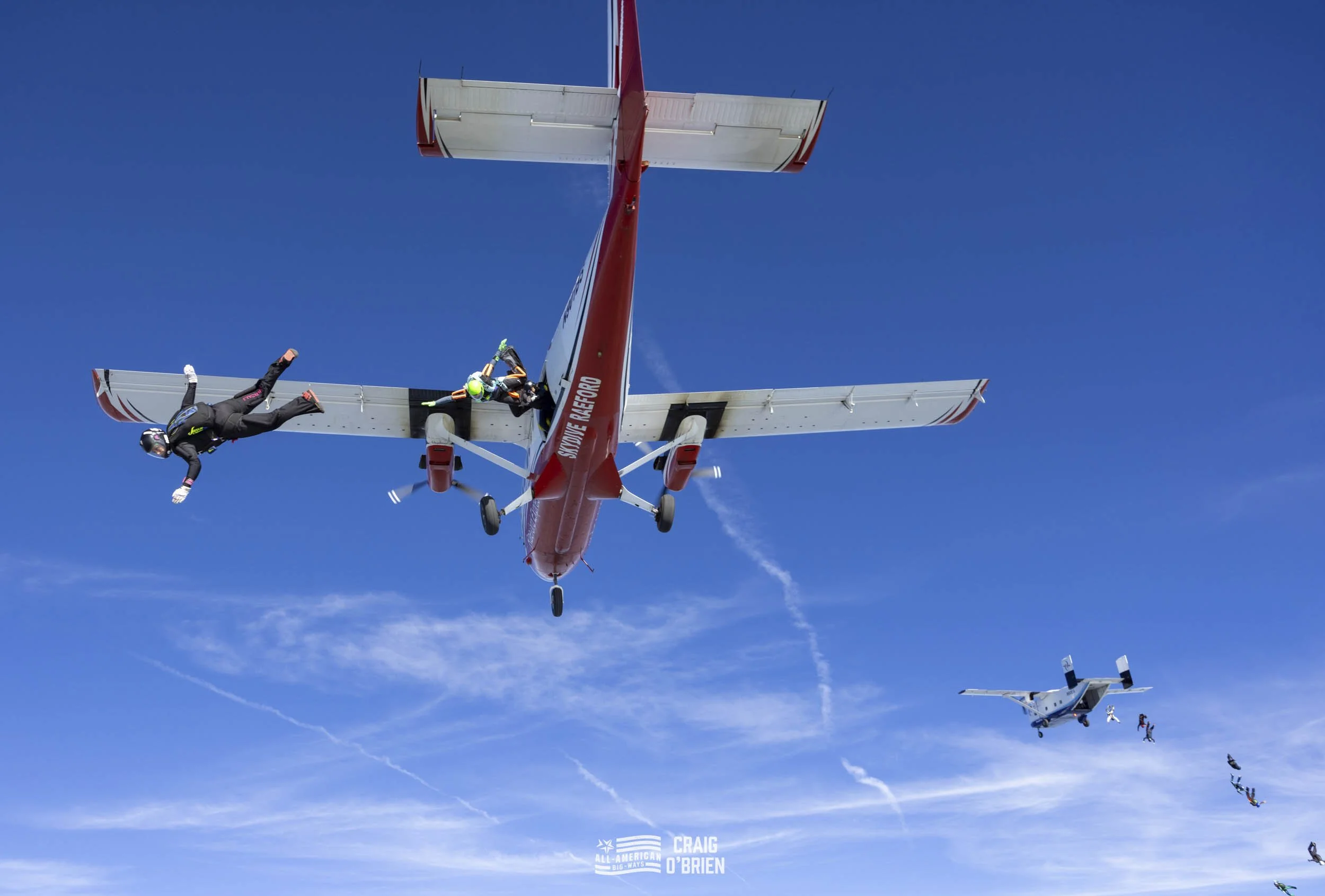 Filming of an aircraft stunt with skydivers falling from different planes against a bright blue sky.