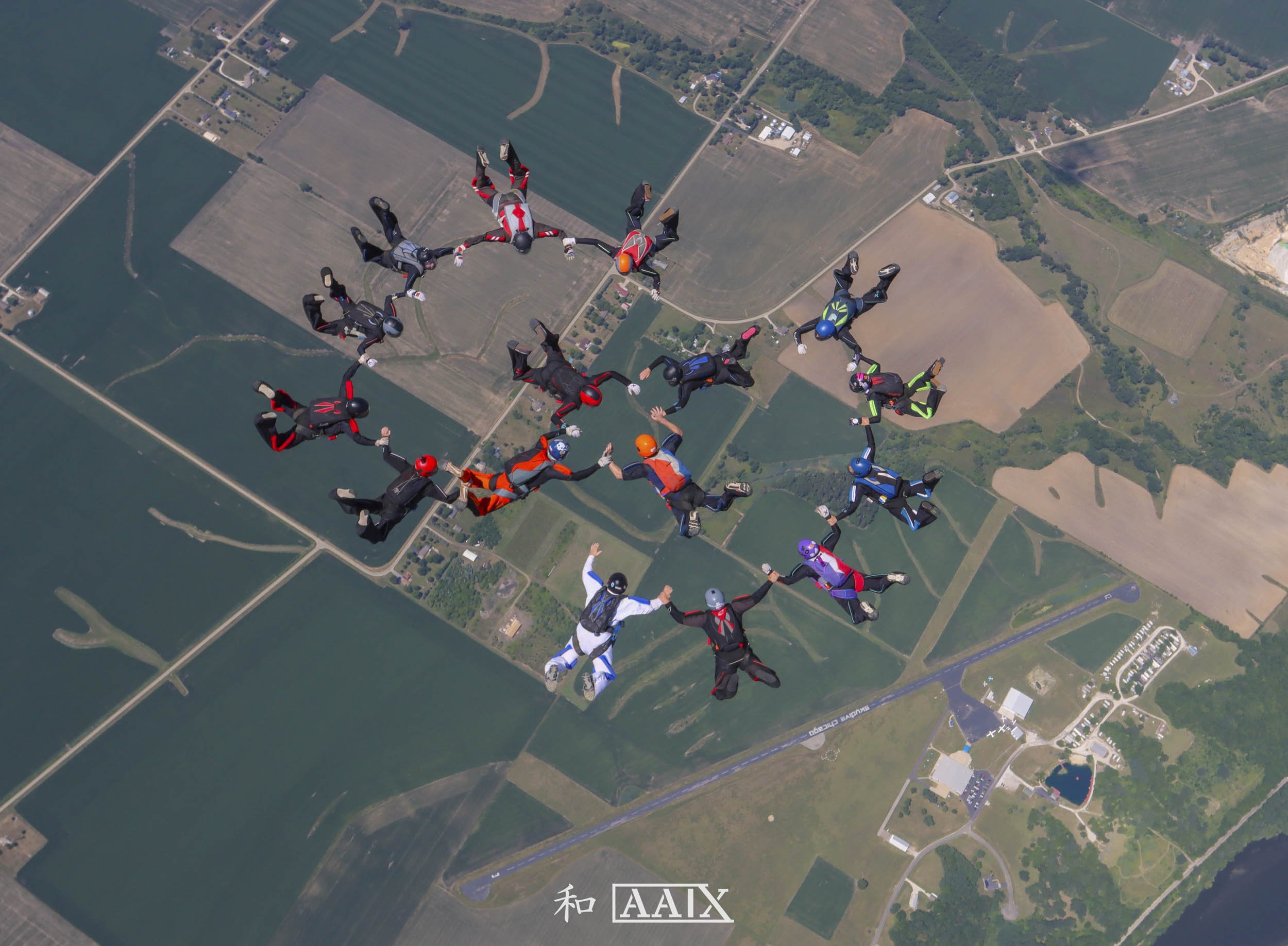 Group of skydivers in jumpsuits and helmets forming a circle while free-falling over a landscape of fields and roads.