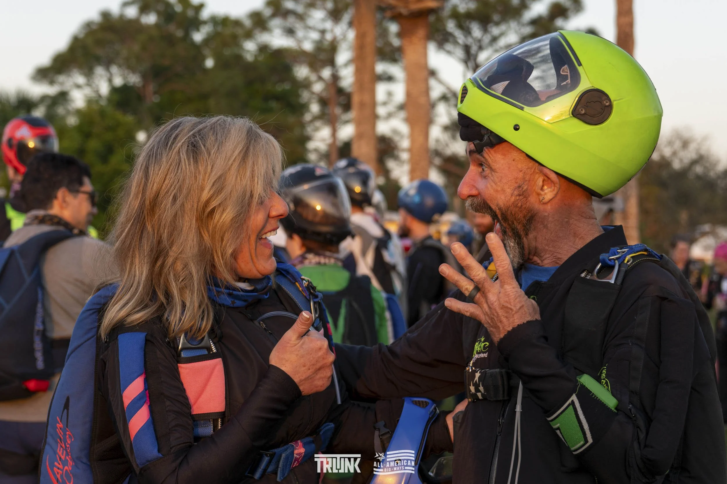 Two motorcyclists, a woman and a man, happily talking together outdoors with trees in the background, surrounded by others wearing helmets.
