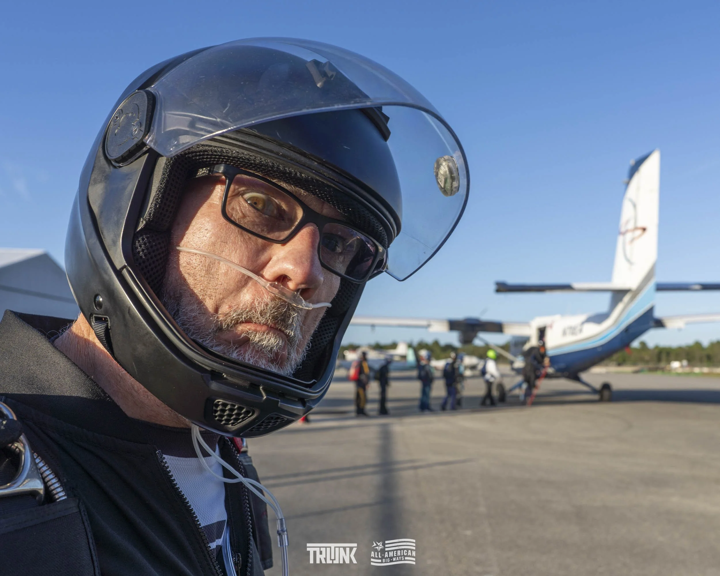 A man in a black motorcycle helmet and glasses with a red nose clip, standing in front of a small airplane on an airport tarmac, with several people in the background near the aircraft.