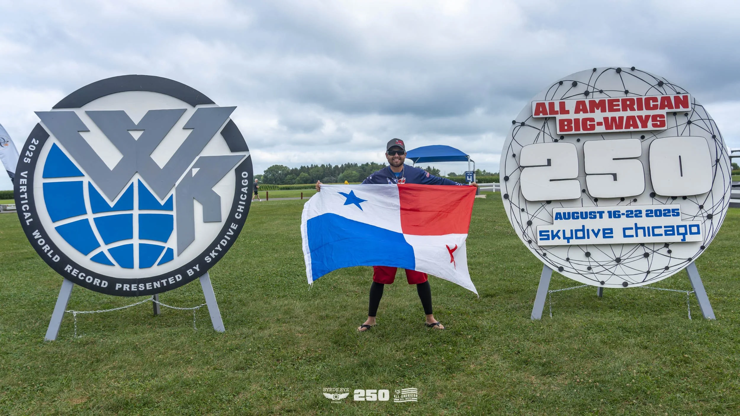 Person holding a Texas flag standing between two large signs at Skydive Chicago event. One sign shows a world record logo and the other indicates 250 big-way jumps from August 16-22, 2025.