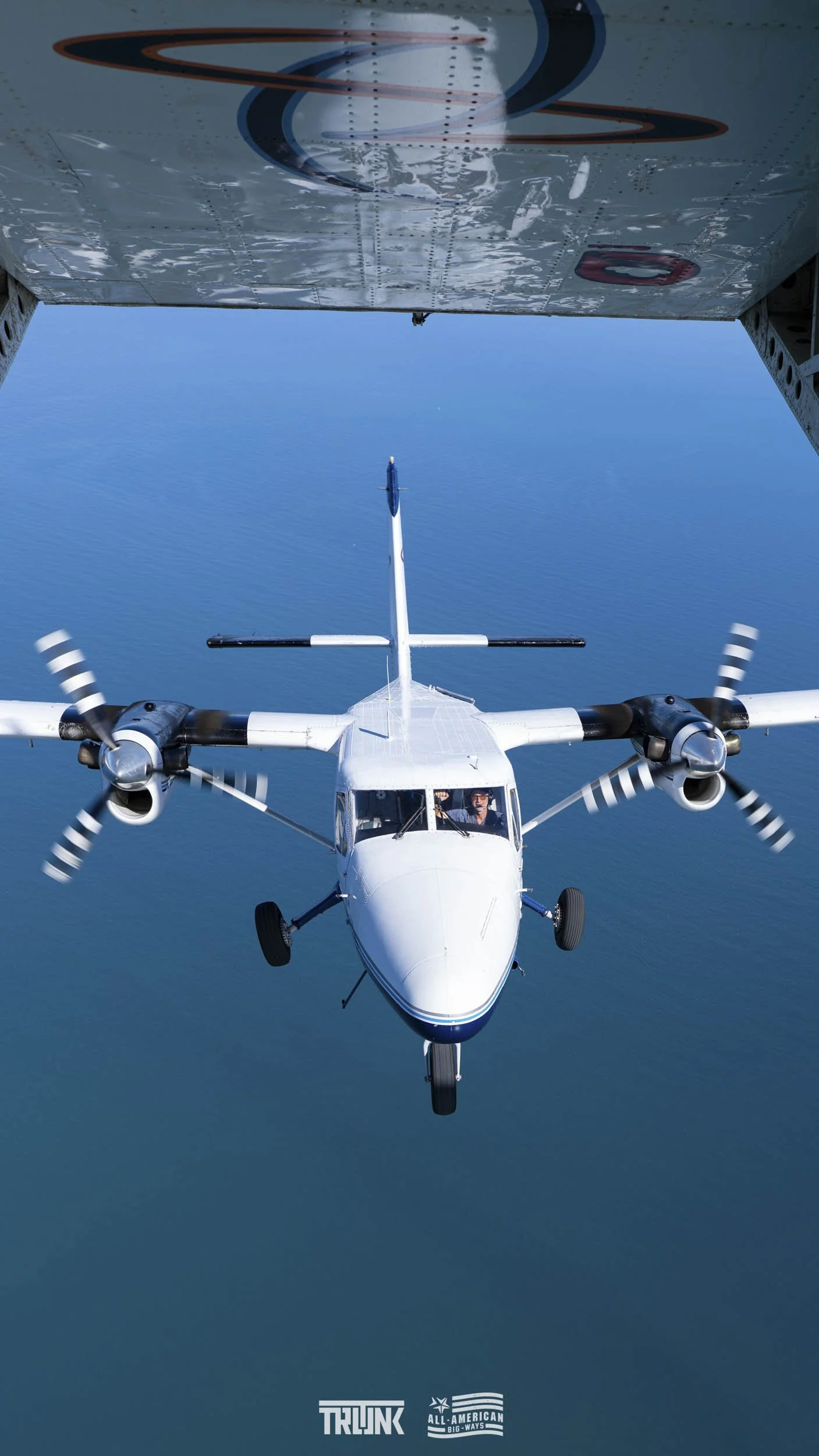A twin-engine aircraft flying above calm blue water, viewed from directly underneath, with another plane's underside visible at the top of the image.