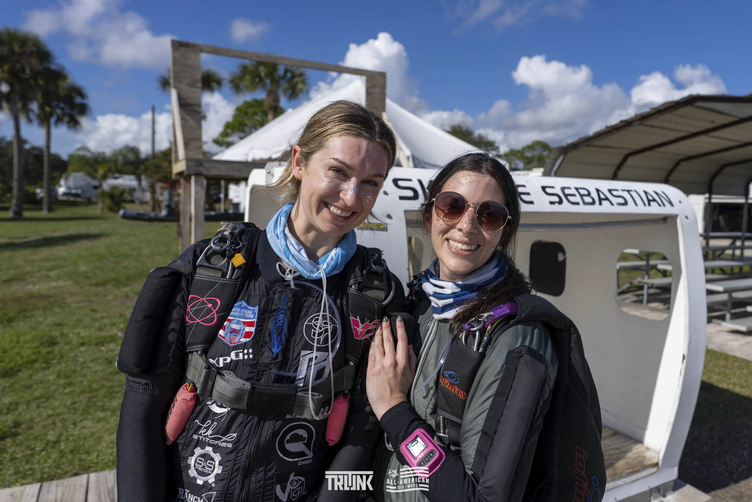 Two female skydivers smiling and posing together at the airport before a jump, with airplane and sky in the background.
